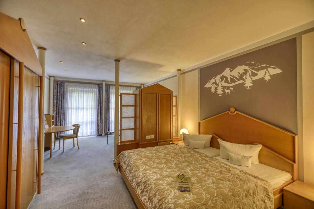 A hotel room with a bed, bedside table, and a wall with a mountain design. There is a desk and chair in the corner, and a door leading to another room.