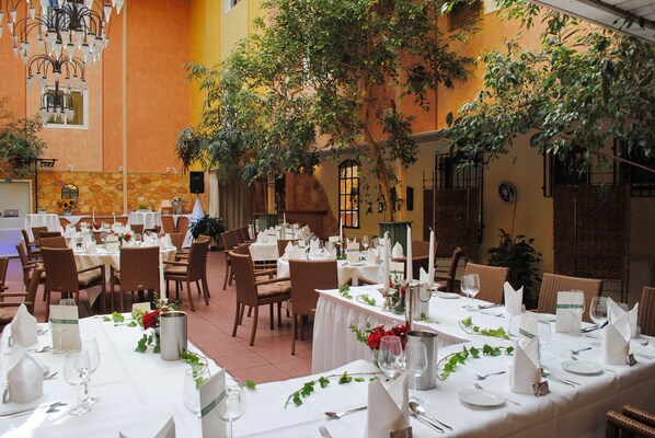 An indoor dining area with multiple tables set for a formal event, surrounded by wicker chairs and potted plants.