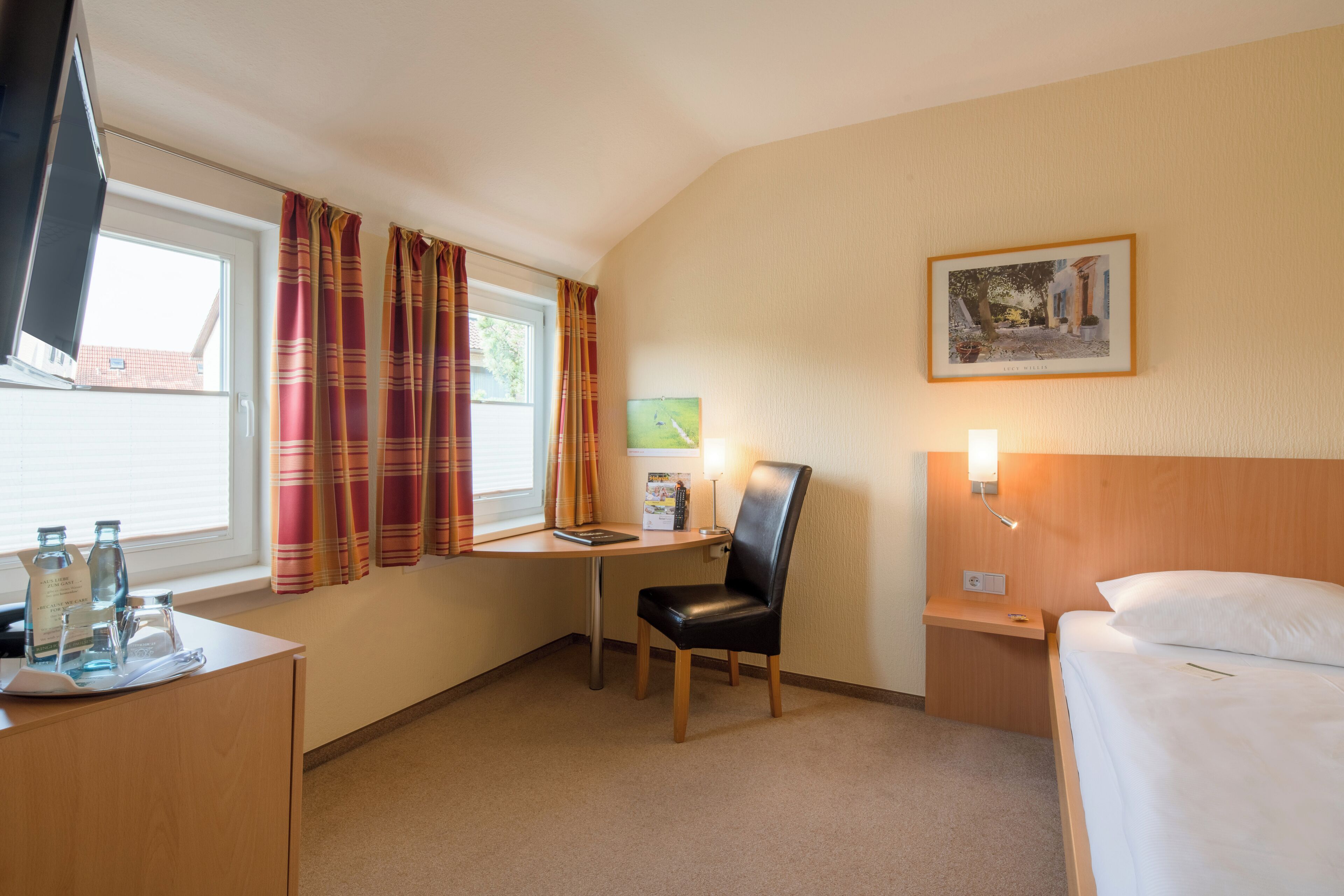 Une chambre d’hôtel comprenant un lit, un bureau avec une chaise, une fenêtre avec des rideaux, un tableau accroché au mur et une table de chevet avec une lampe.