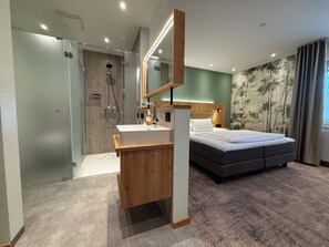 A modern hotel room with a wooden vanity, a large bed, a shower, and a wall-mounted mirror. - Ringhotel Paulsen (Zeven)
