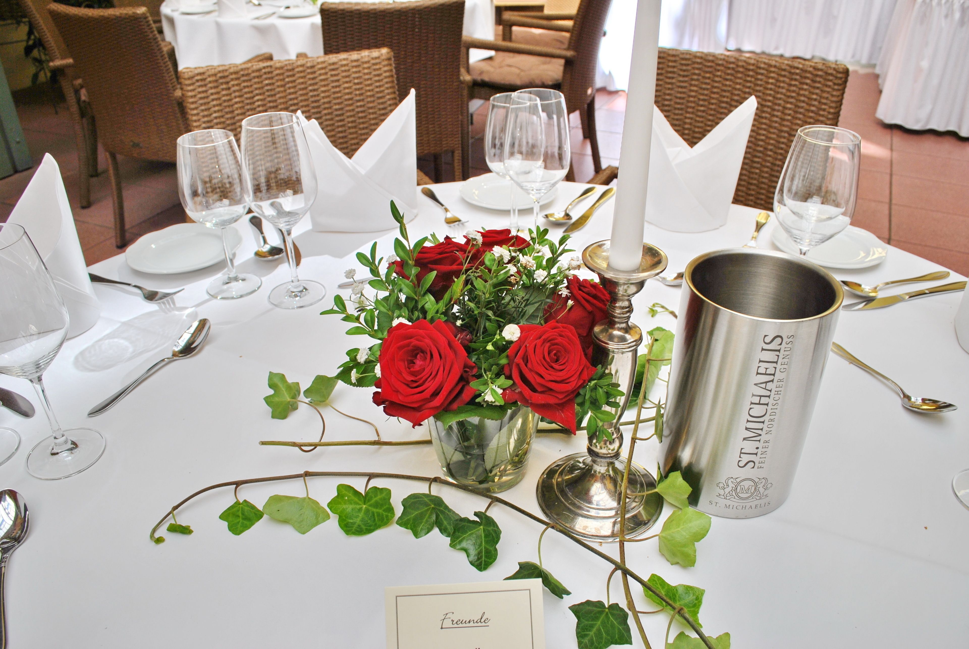 A dining table set with white tablecloths, red roses, and a St. Michaelis bottle.