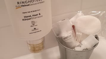 A bathroom with a bottle of hand, hair, and body wash, a shower cap, and a metal bucket containing toiletries.