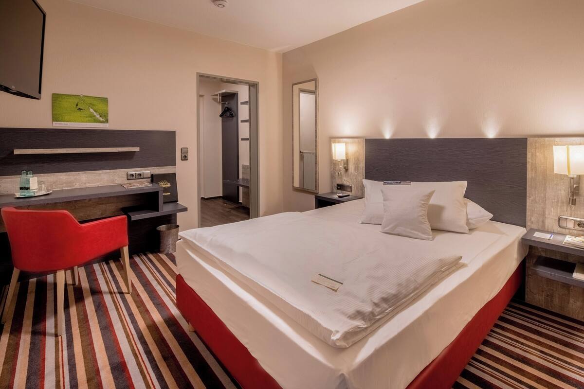 Habitación de hotel con una cama grande, un escritorio con silla, un espejo y televisor montado en la pared.