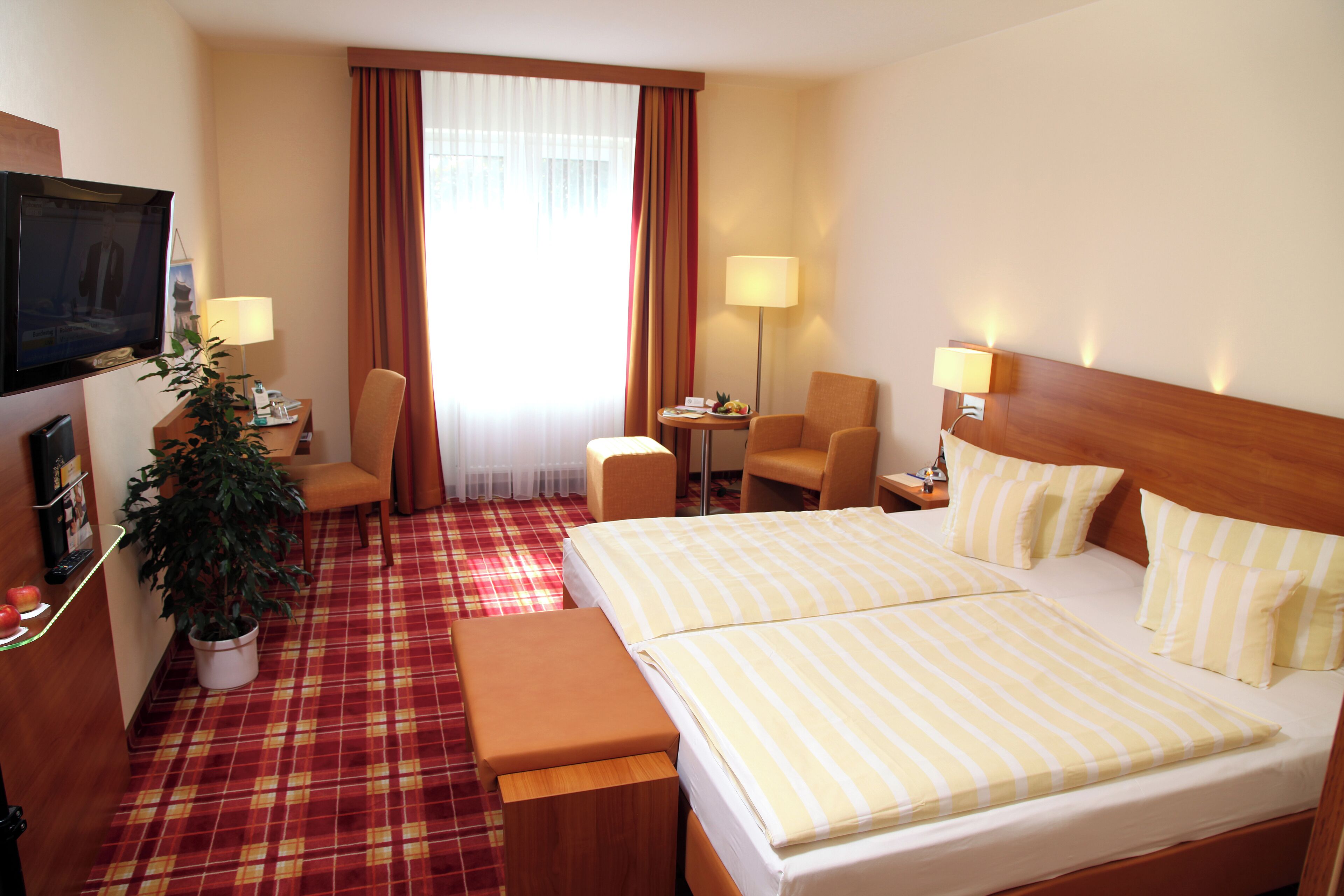 A hotel room with a bed, two chairs, a desk, a TV, and a potted plant.