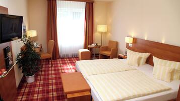 A hotel room with a bed, two chairs, a desk, a TV, and a potted plant.