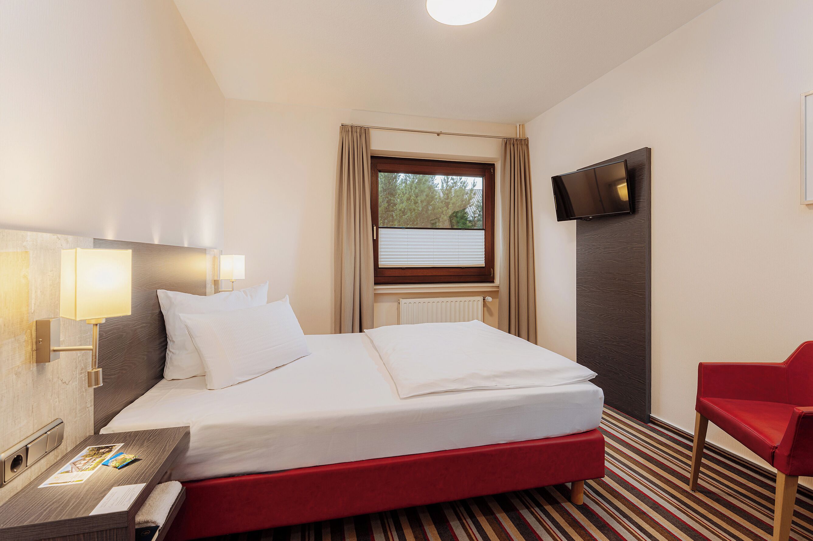 Ein Hotelzimmer mit einem Bett, einem Schreibtisch mit Lampe, einem an der Wand befestigten Fernseher und einem Fenster mit Vorhängen.
