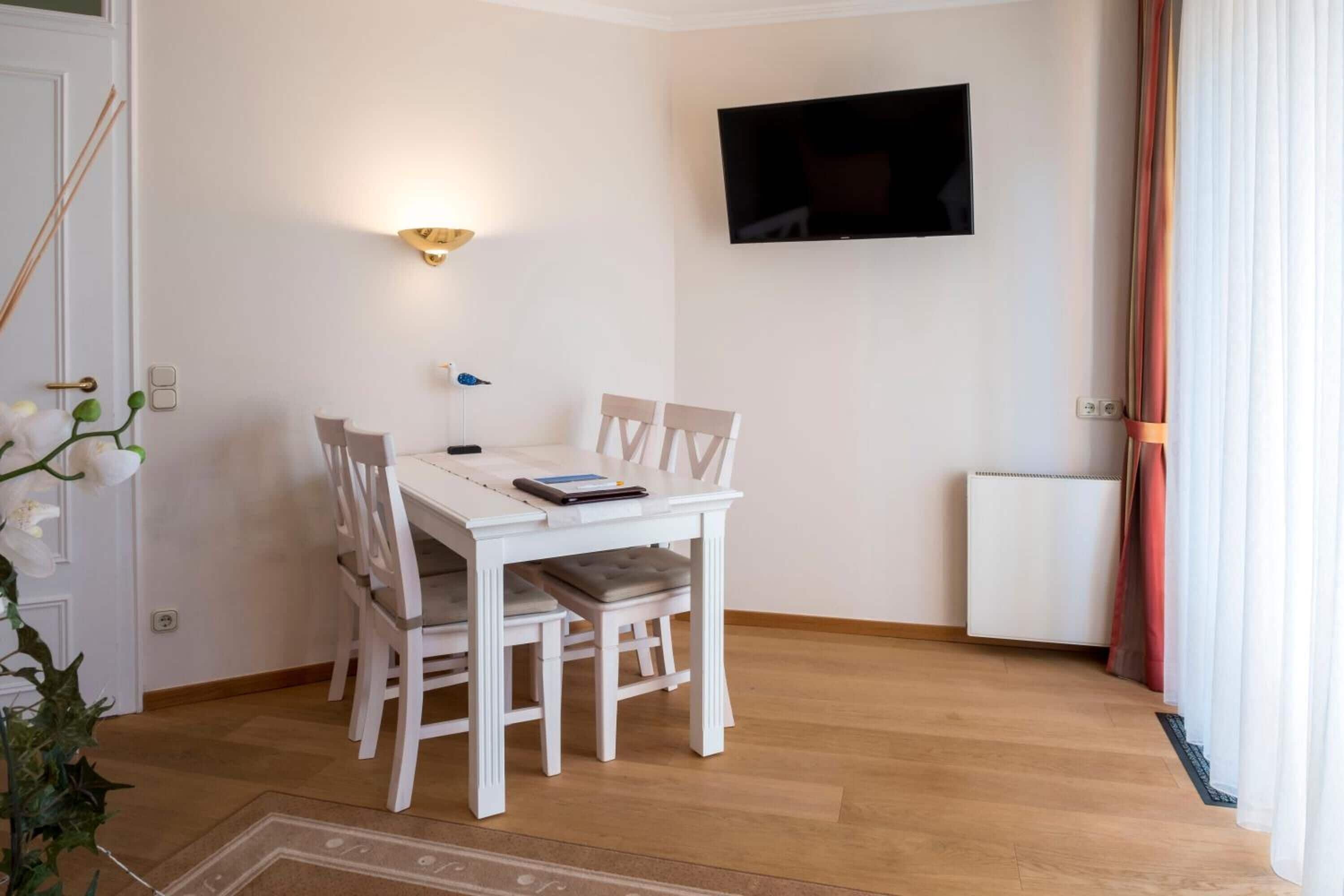 Ein Essbereich mit weißem Tisch und Stühlen, einem an der Wand befestigten Fernseher und einer Heizung.