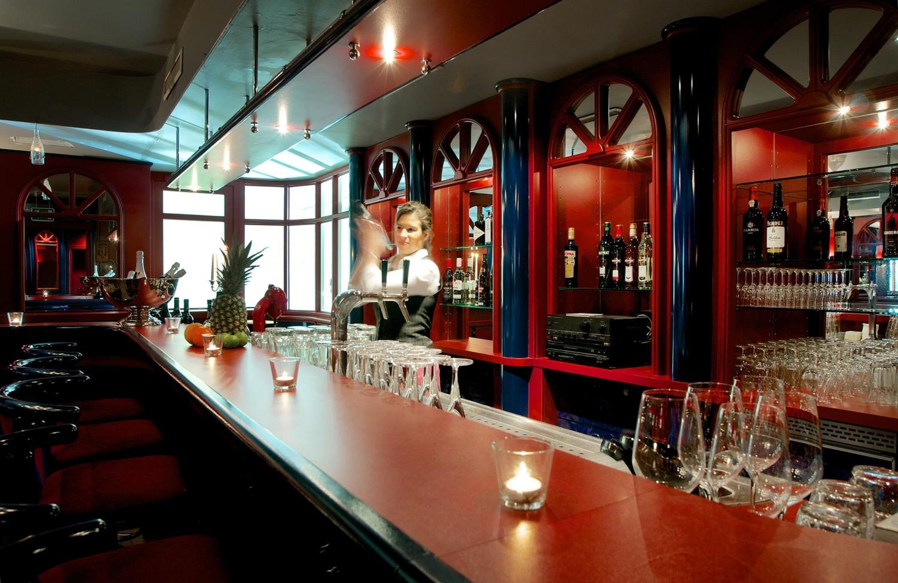 Uma área de bar com um bartender, um balcão comprido com copos e garrafas, e uma grande janela ao fundo.