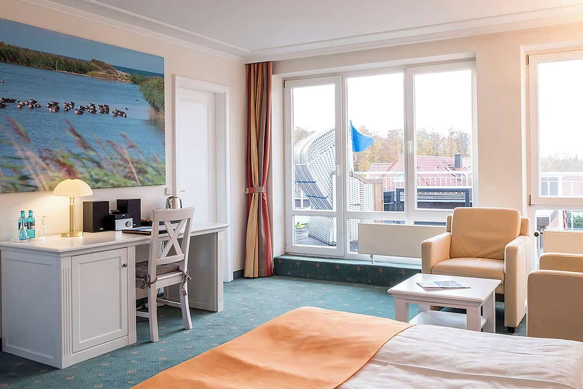A hotel room with a bed, a desk, a chair, a small table, and a large window with a view of buildings and a flag.