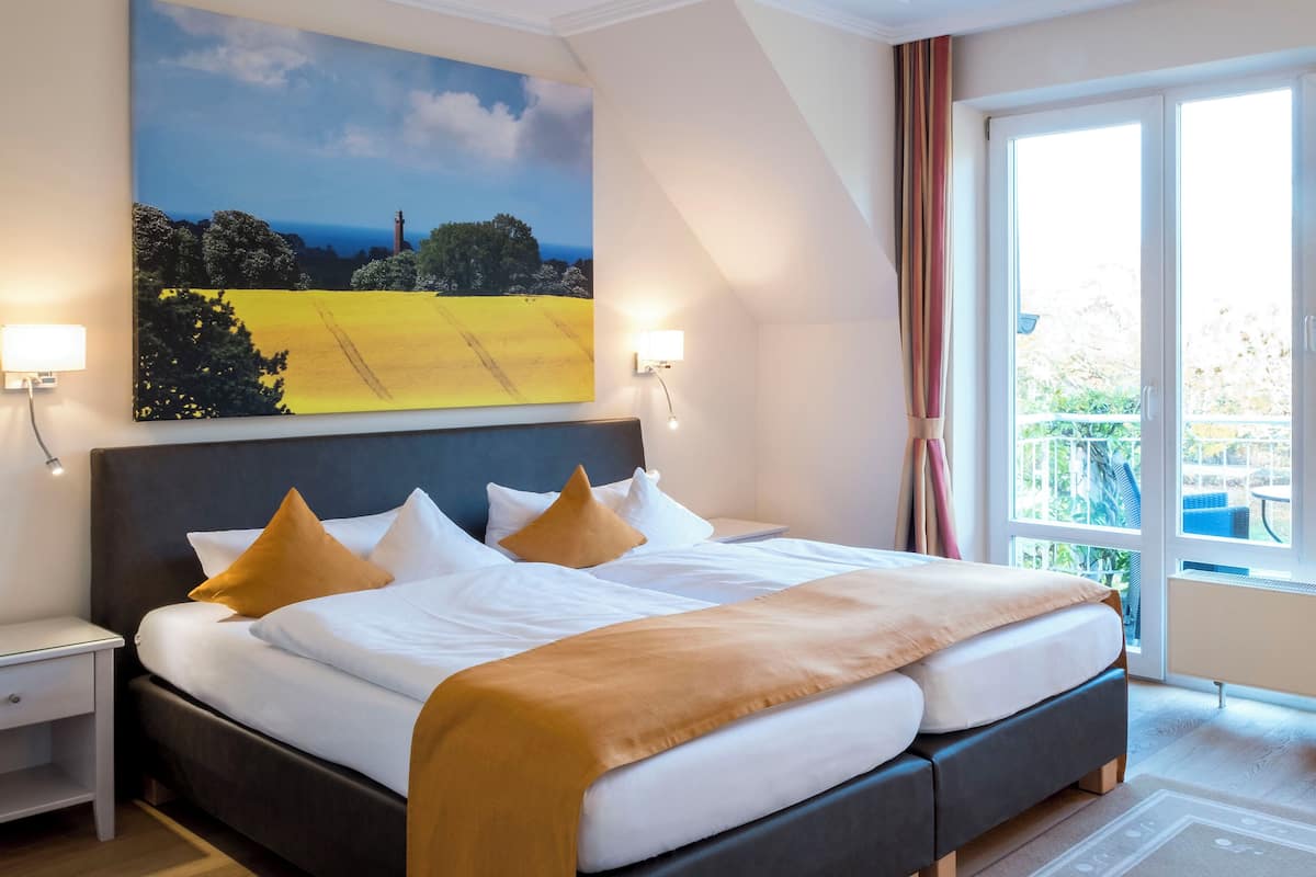 A hotel room with a large bed, a nightstand, a lamp, a painting of a landscape, and a view of greenery through the window.