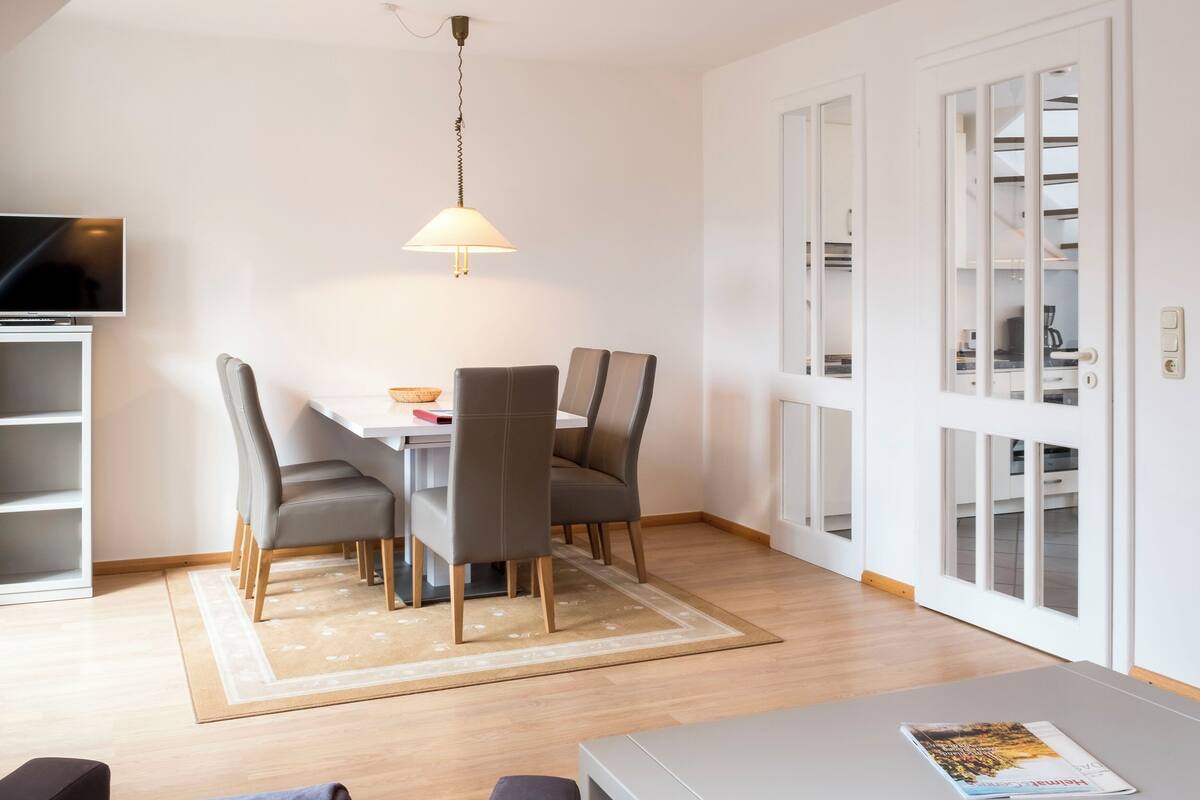 A modern dining area with a table, chairs, and a lamp. There is a TV mounted on the wall and a door with glass panels.