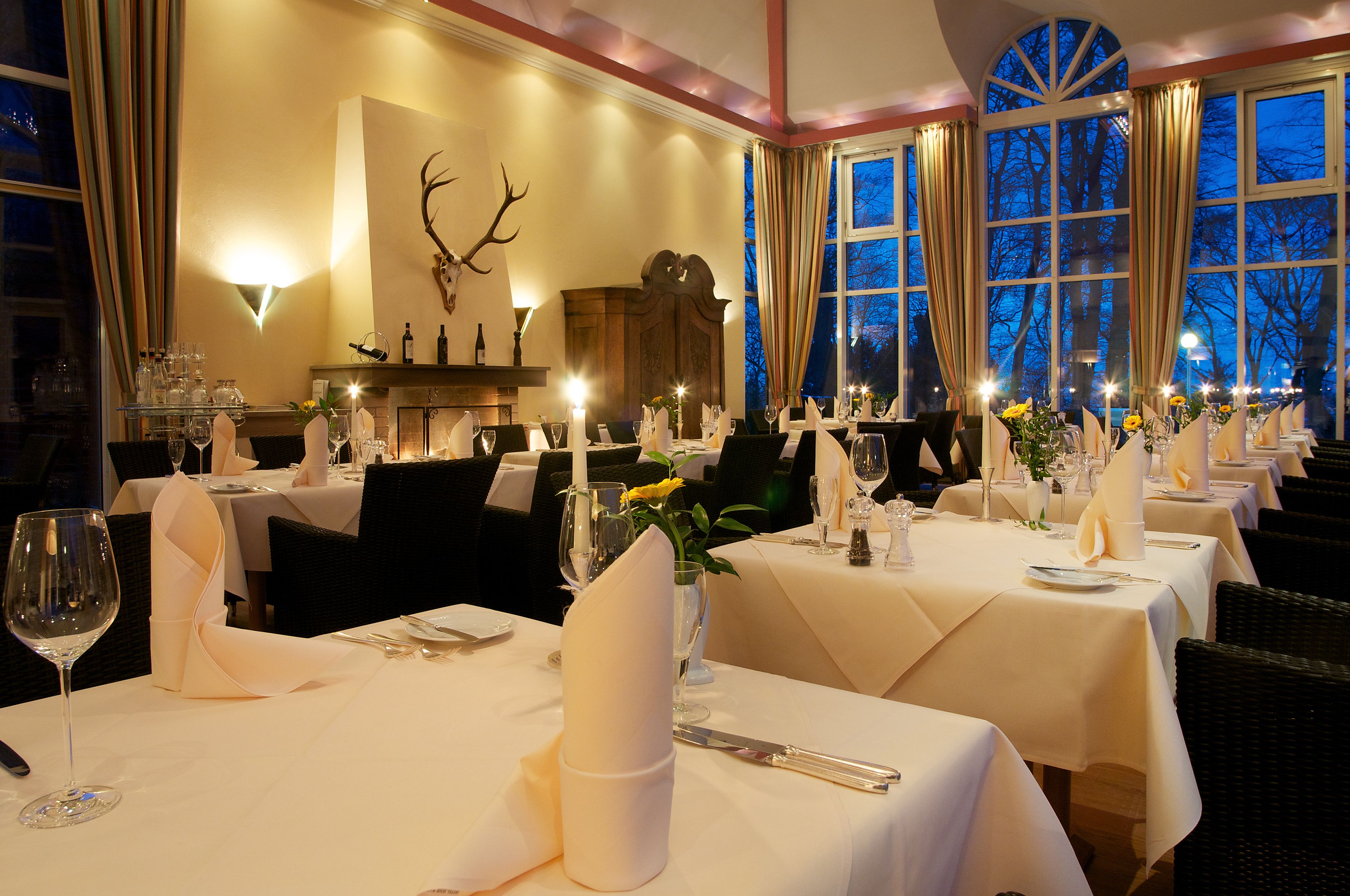 A formal dining room with tables set for a meal, featuring candles, wine glasses, and floral centerpieces.