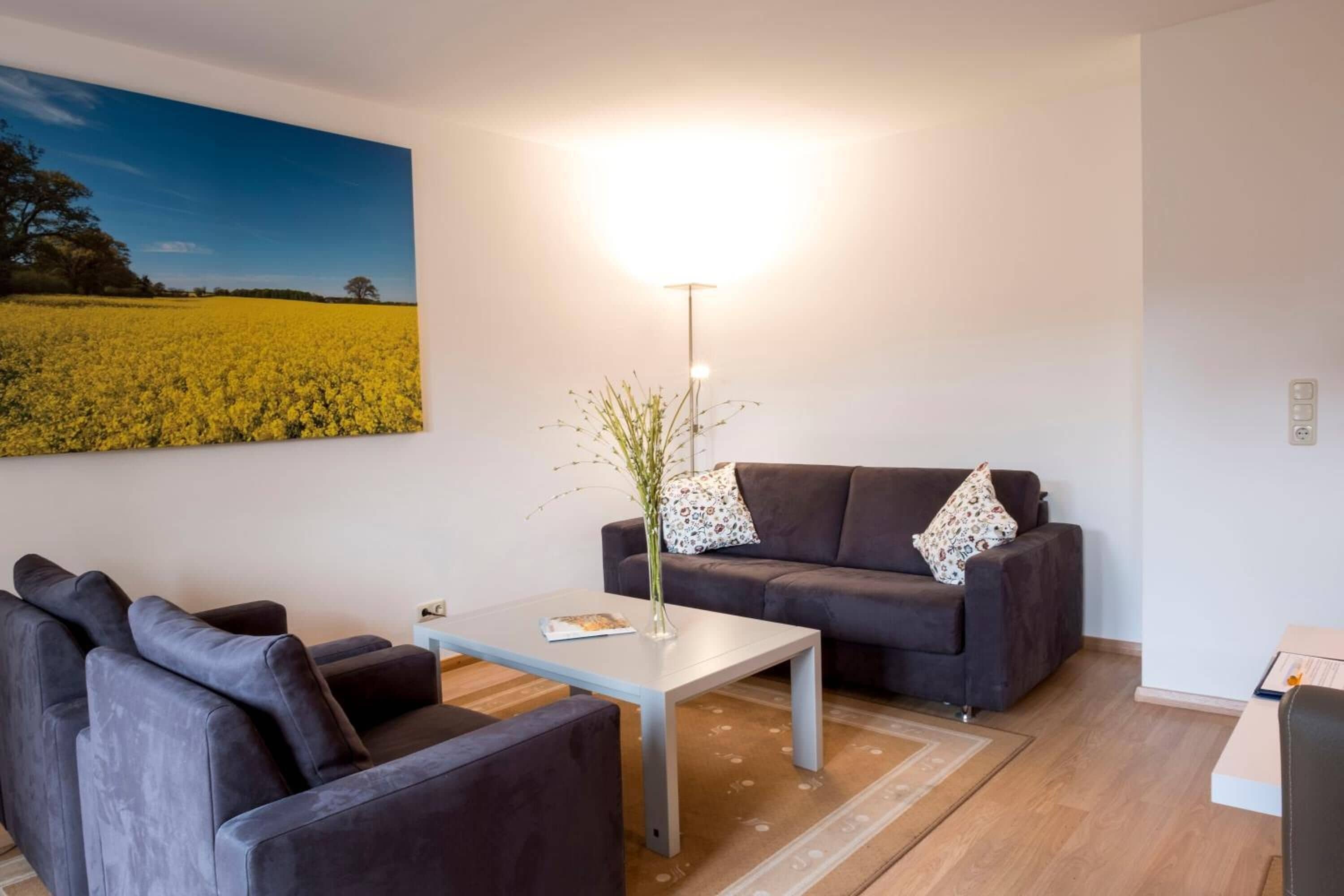 Una sala de estar moderna con un sofá, un sillón y una mesa de centro. Un cuadro de un campo está colgado en la pared, y hay una lámpara de pie sobre la mesa.