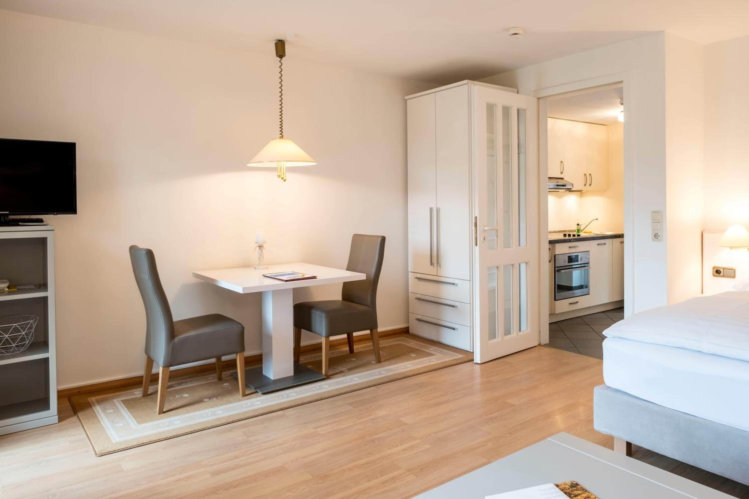 A hotel room with a bed, a TV, a dining table with chairs, a kitchen area, and a wardrobe.