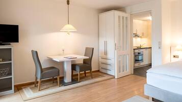 A hotel room with a bed, a TV, a dining table with chairs, a kitchen area, and a wardrobe.