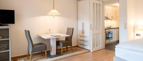 A hotel room with a bed, a TV, a dining table with chairs, a kitchen area, and a wardrobe.