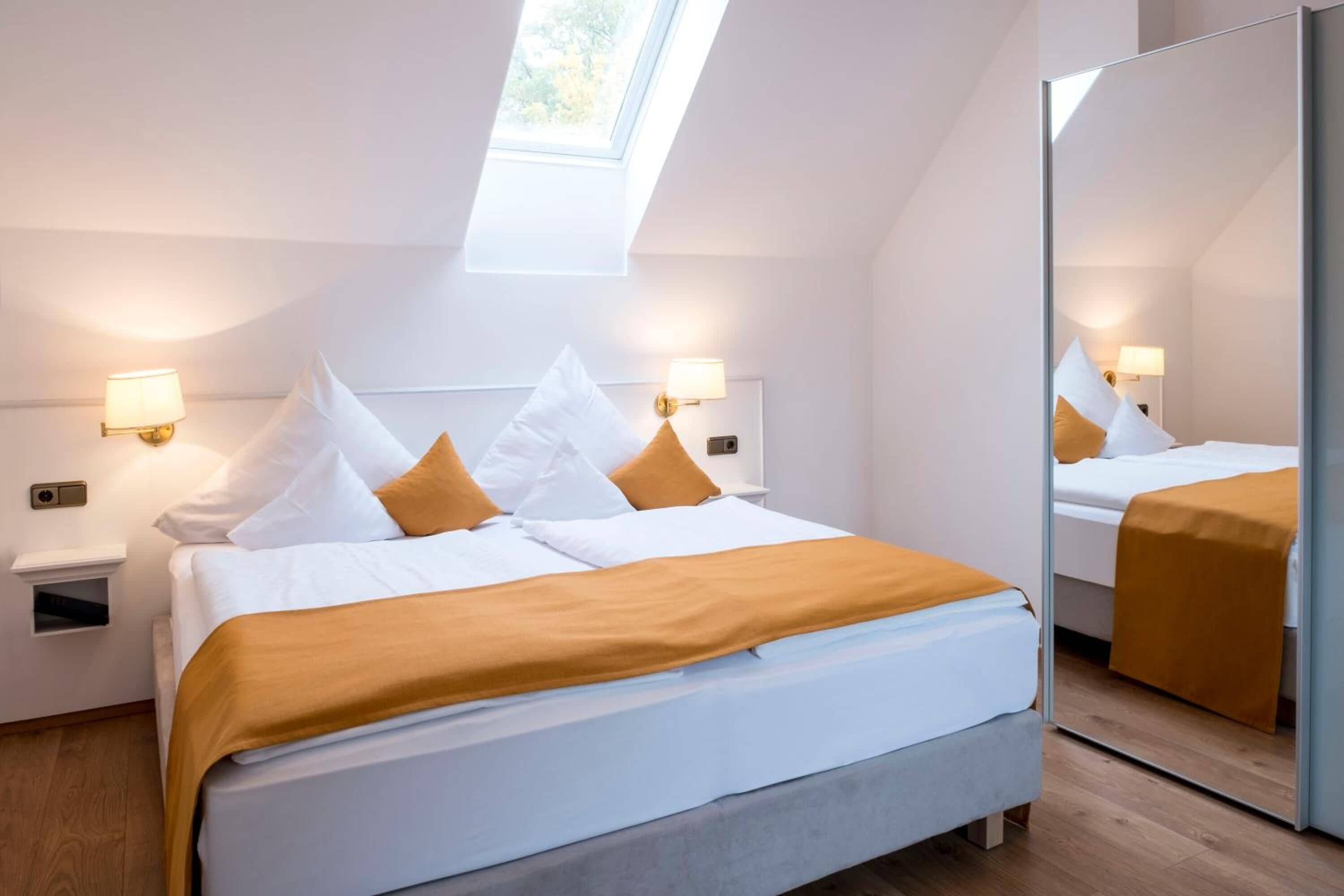 A modern hotel room with a skylight, a large bed with white bedding and orange accents, a bedside table with a lamp, and a full-length mirror.