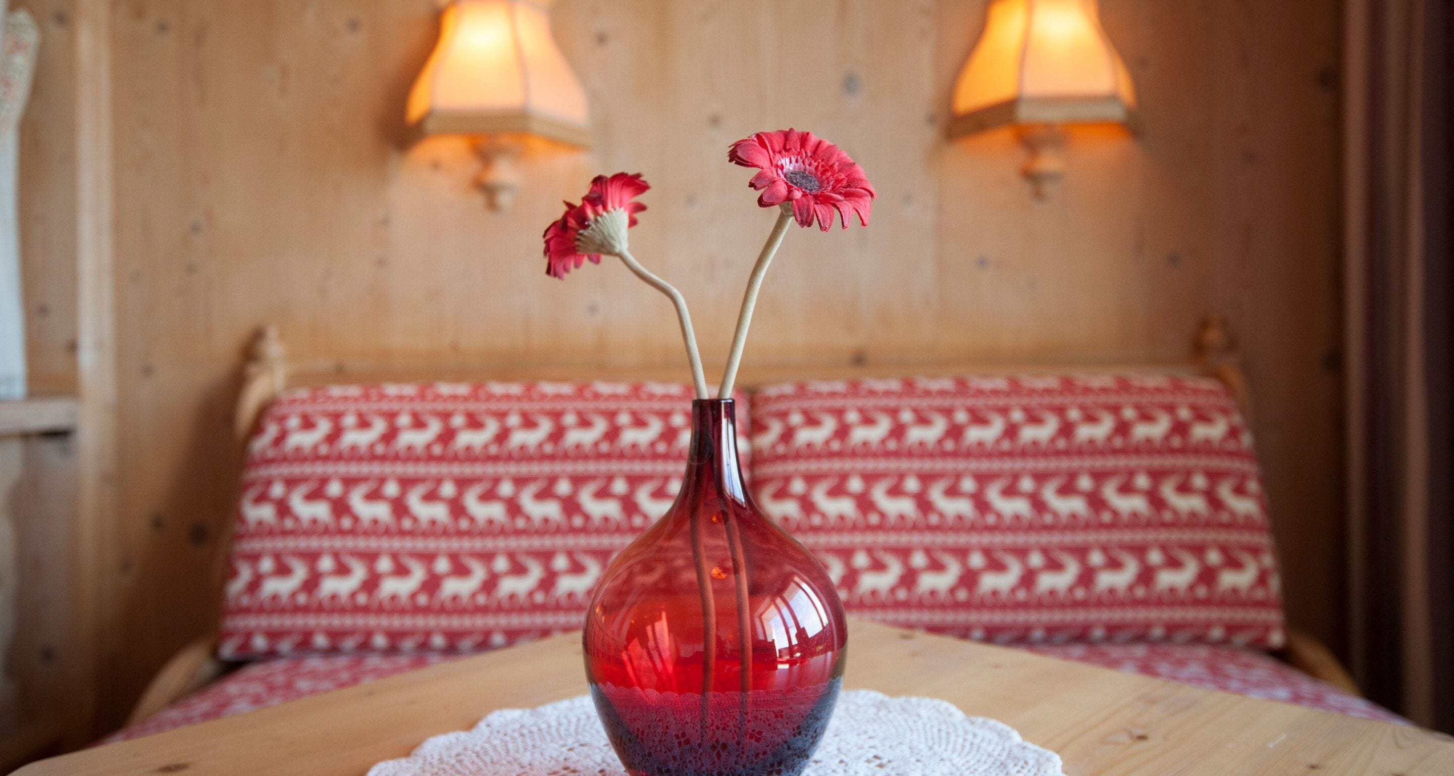 Un vase rouge orné de deux fleurs roses, posé sur une table en bois dans une pièce aux murs à motifs et éclairée par deux lampes fixées au mur.