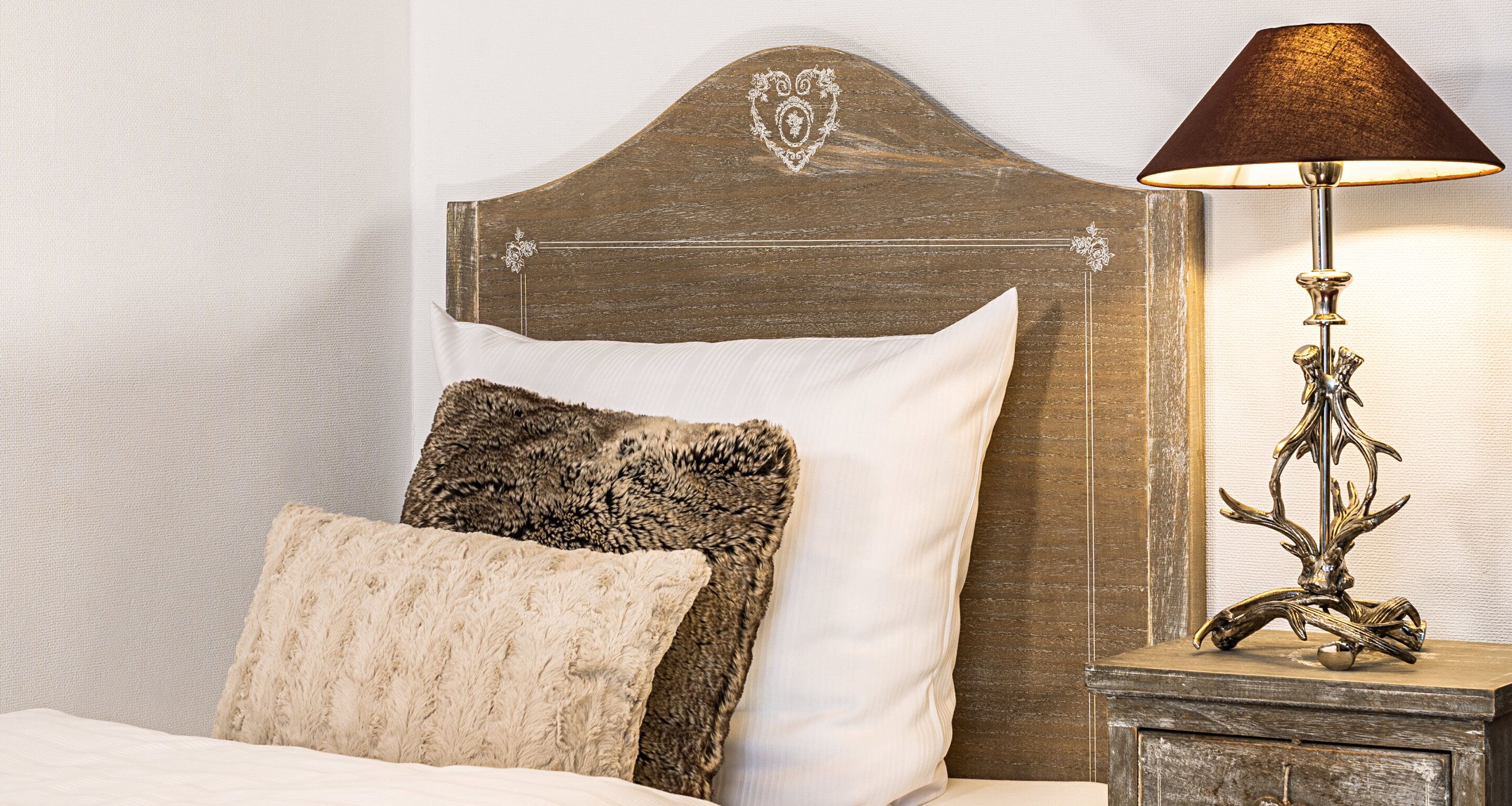 Una cama con cabecera de madera, un almohadón beige suave, un plaid de piel sintética marrón oscuro y una lámpara decorativa sobre una mesita de noche de madera.