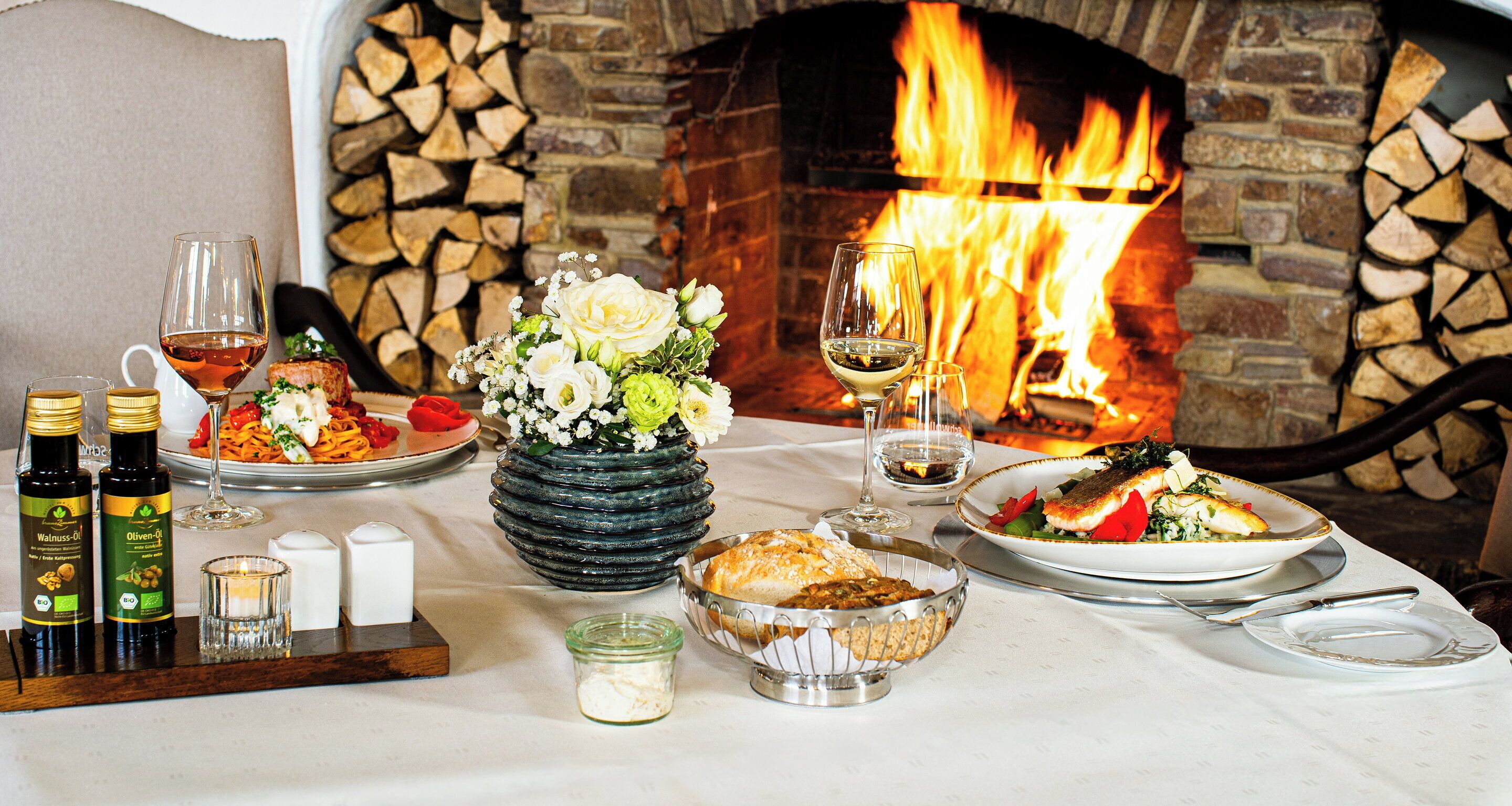 Une table à manger dressée avec des assiettes remplies de nourriture, des verres à vin et des bouteilles d’huile d’olive, le tout placé devant une cheminée.