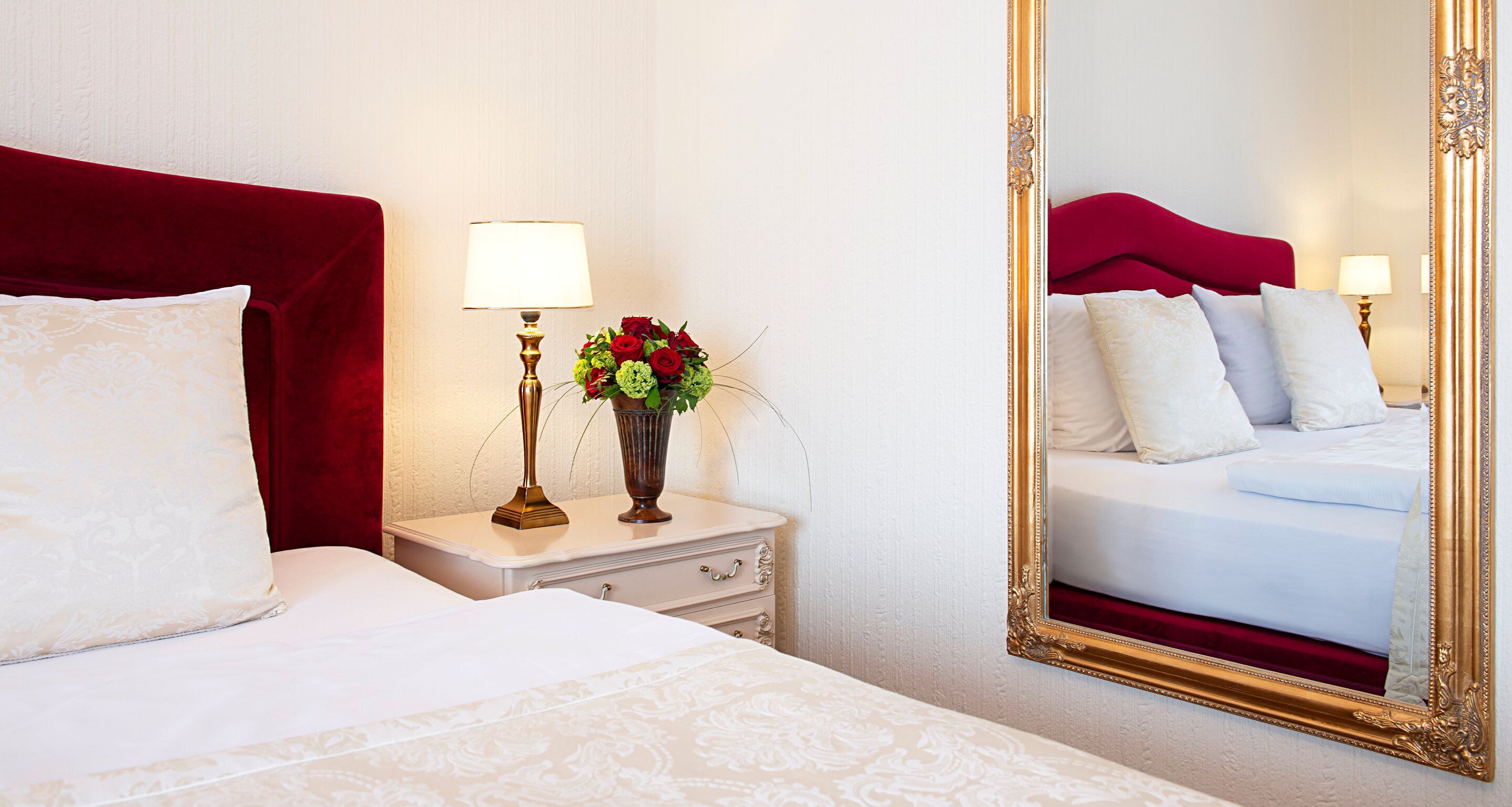 Une chambre d’hôtel avec un grand lit, une table de chevet avec une lampe et un vase de fleurs, et un miroir avec un cadre doré.