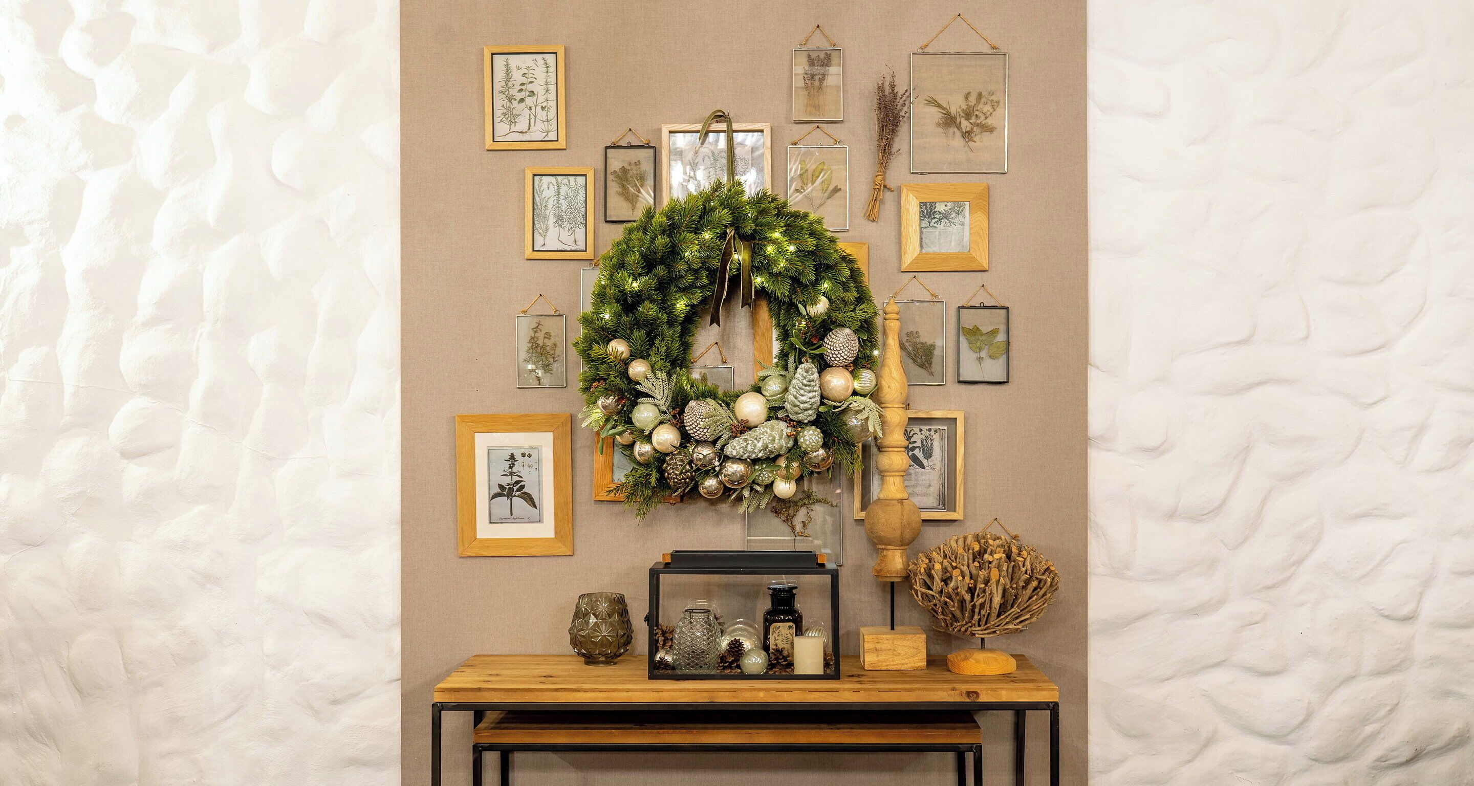 A wall with framed botanical prints, a wreath with pine cones, and a wooden table with a candle and a glass jar.