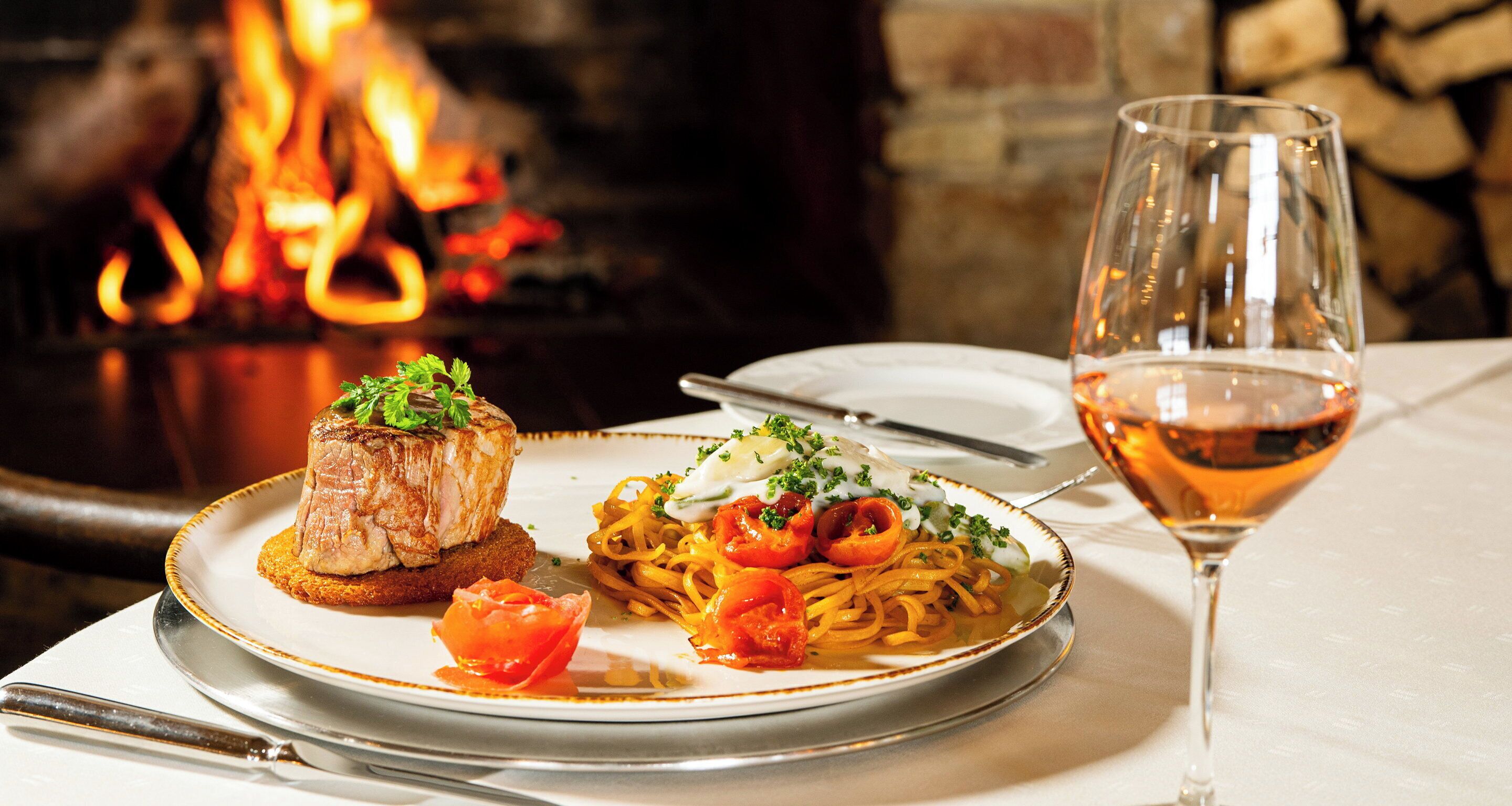 A plate of food with a glass of wine and a fire in the background.