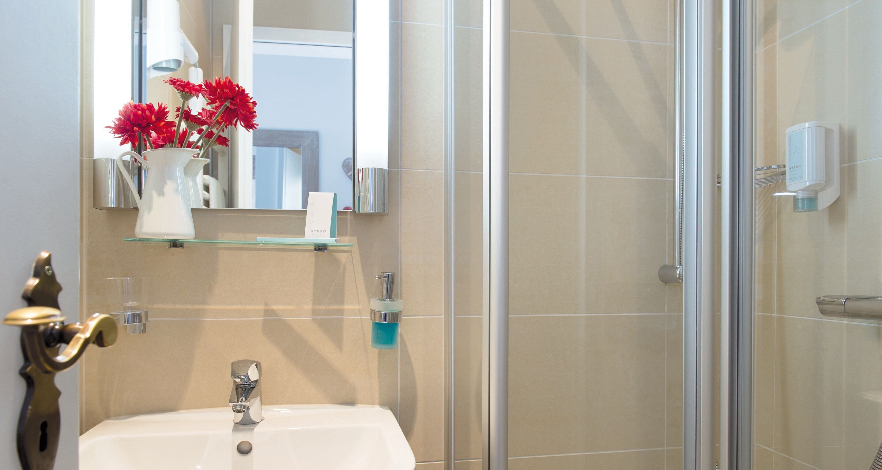 A bathroom with a white sink, a mirror, a vase with red flowers, a glass shelf, and a shower area.