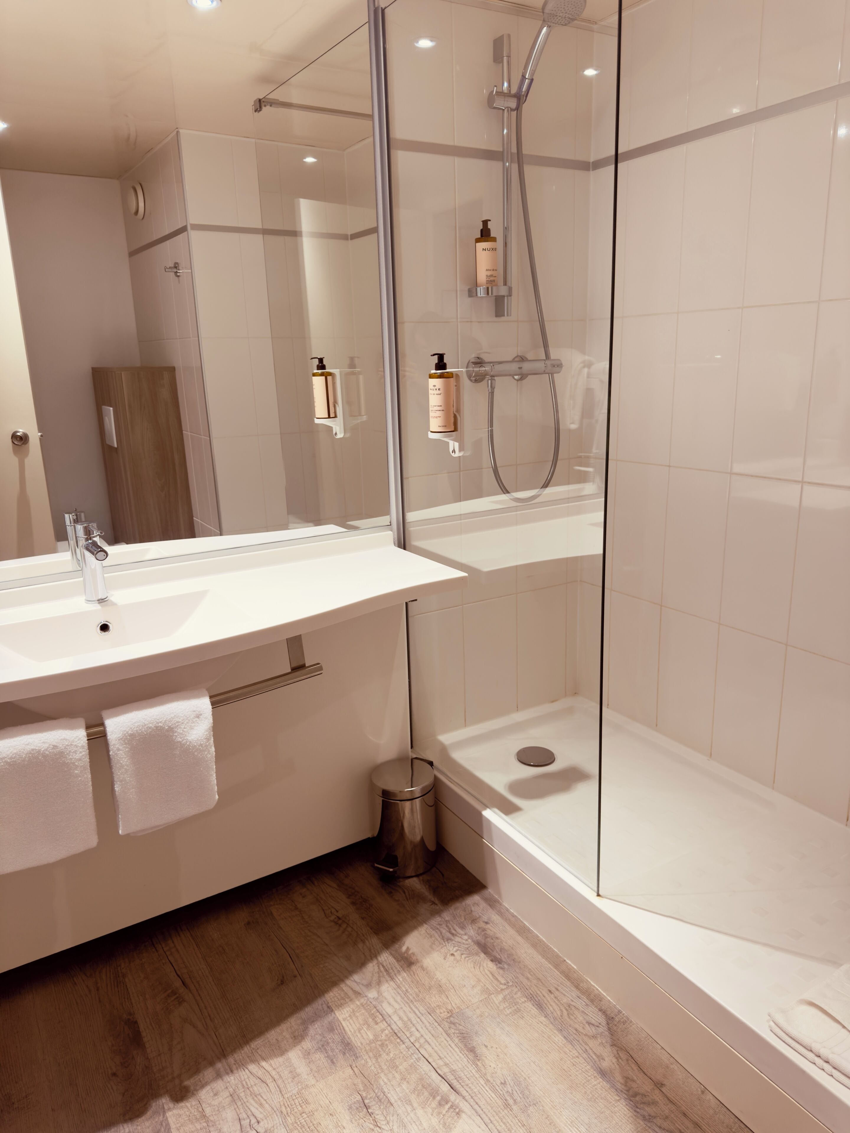 A modern bathroom with a glass-enclosed shower, a large mirror, and a wooden floor.