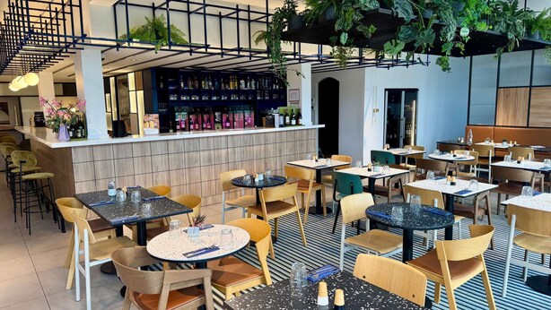 A modern restaurant interior with multiple tables and chairs, a bar area with shelves of bottles, and hanging plants. - Mercure Rouen Val De Reuil (Val-de-Reuil)