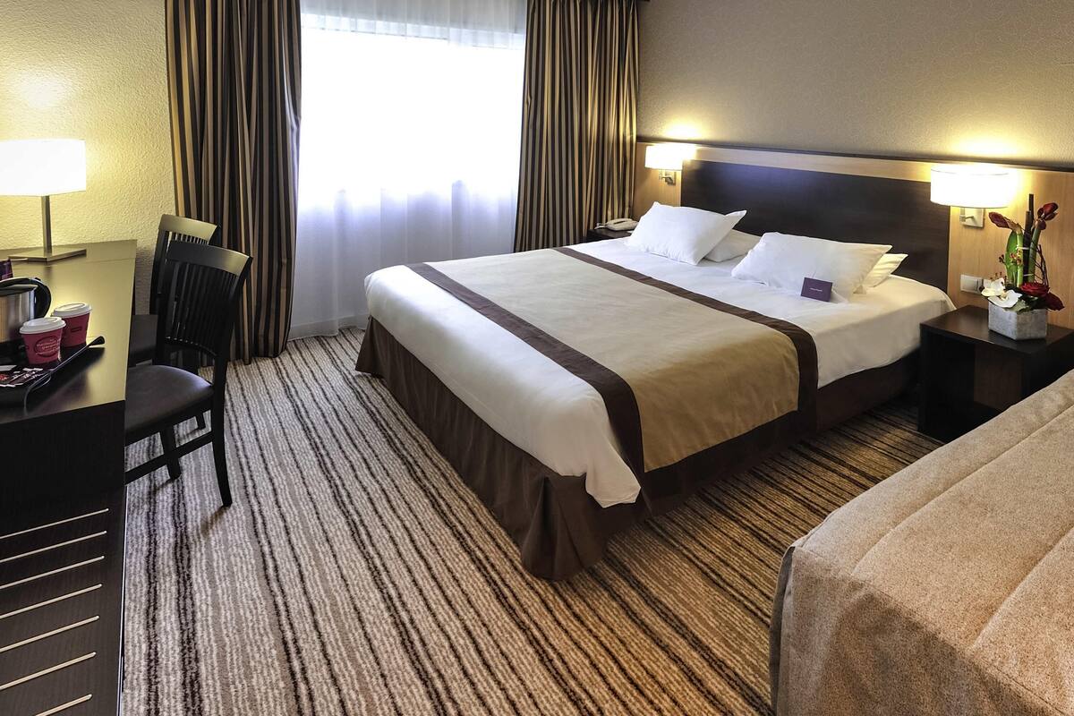 A hotel room with a large bed, a desk with a chair, a nightstand with a lamp, and a window with curtains.