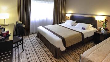 A hotel room with a large bed, a desk with a chair, a nightstand with a lamp, and a window with curtains.