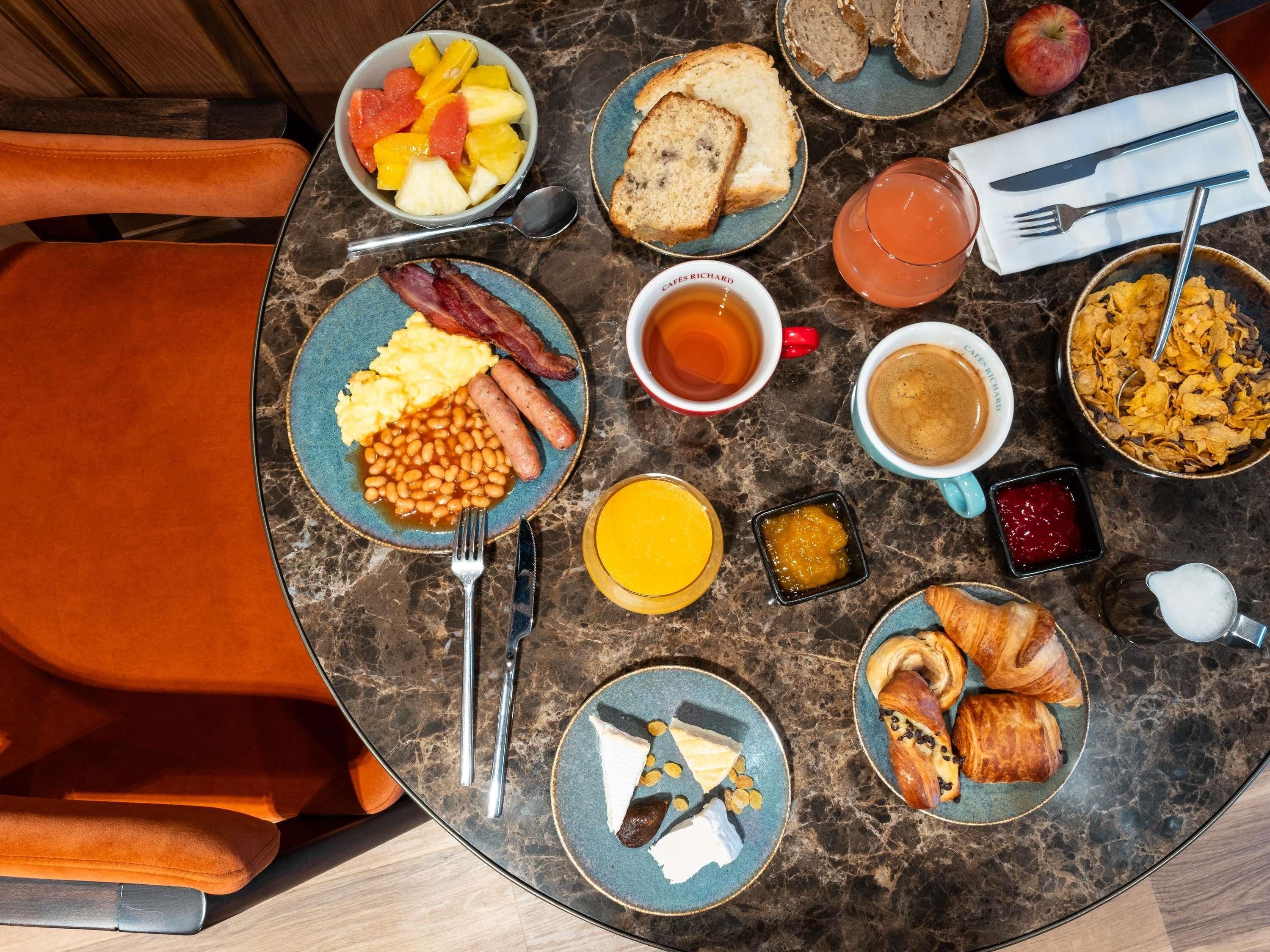 A breakfast table with plates of eggs, bacon, beans, toast, fruit, and pastries.