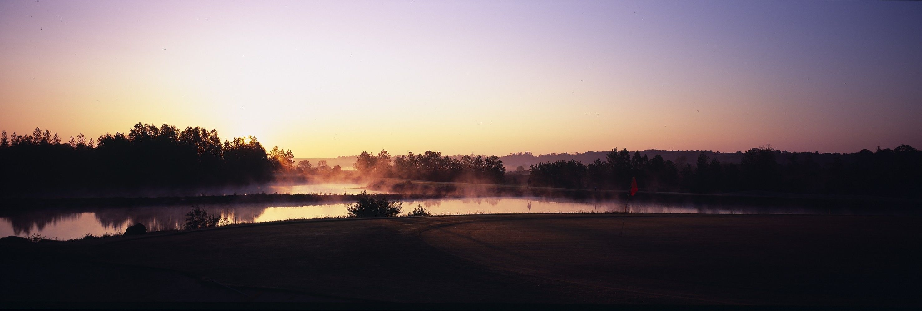 A serene lakeside view at sunrise with a misty atmosphere and a distant flag.