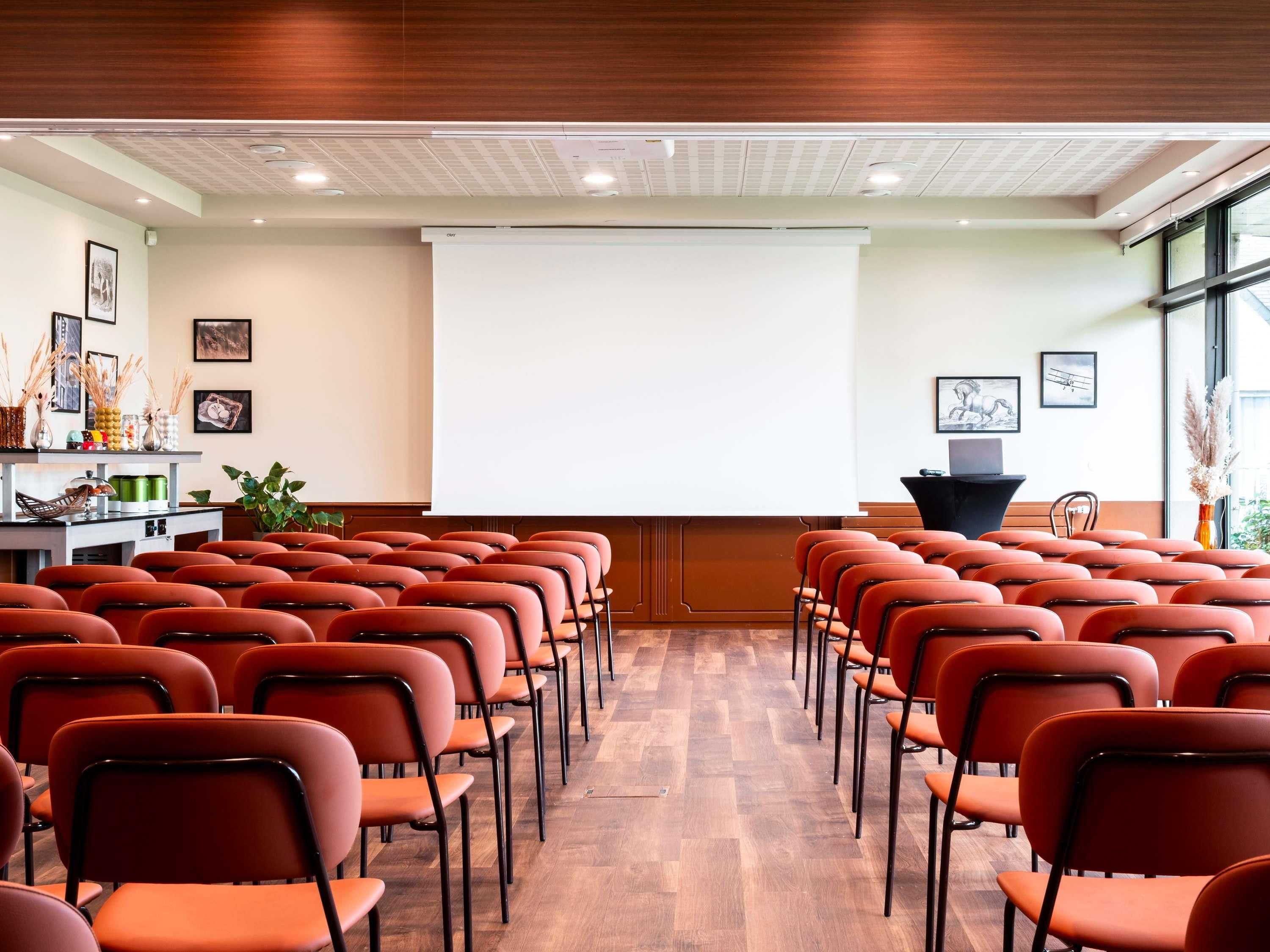 Une salle de conférence avec des rangées de chaises orange, un écran de projection et un pupitre.