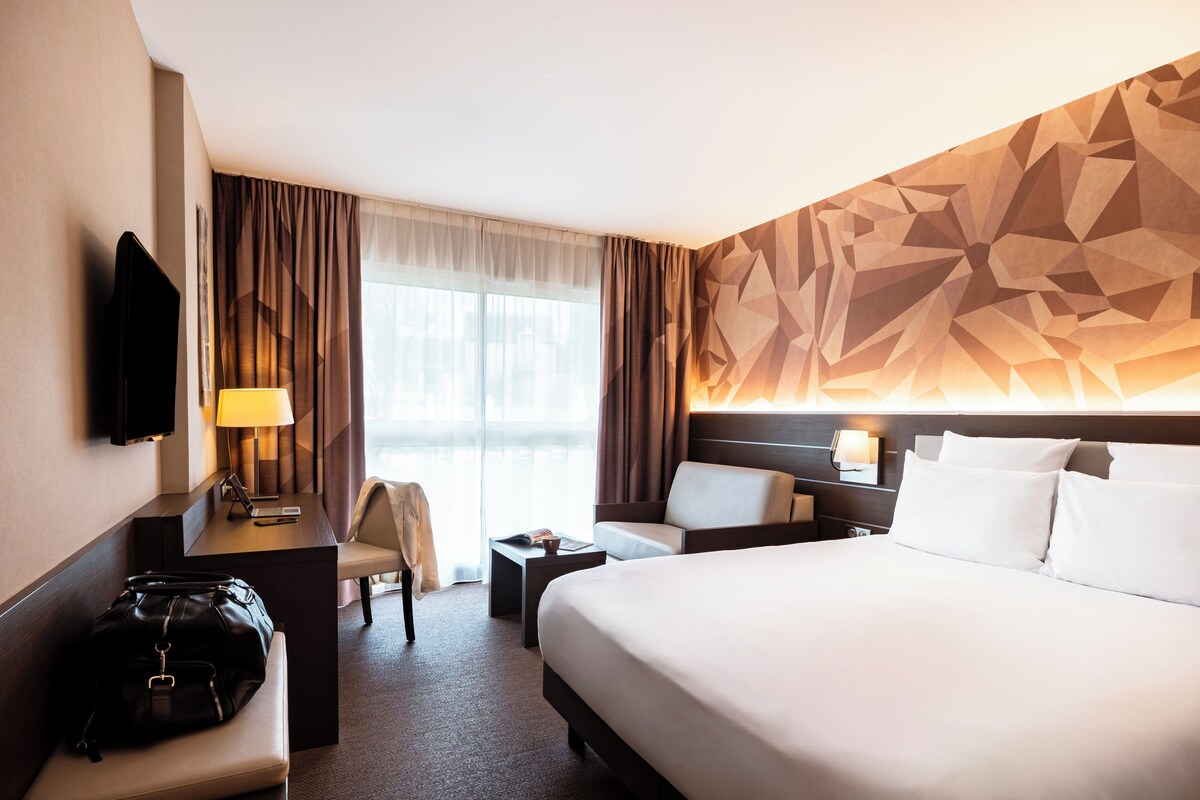 A hotel room with a large bed, a desk with a chair, a TV, and a wall with geometric patterns.