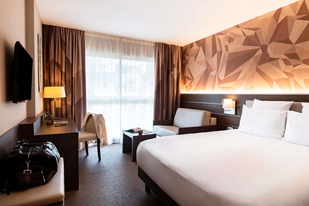 A hotel room with a bed, a desk with a lamp, a chair, a TV, and a large window with curtains.