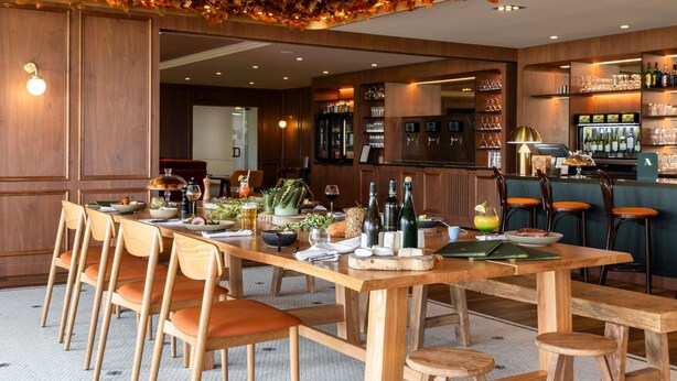 Un restaurante con una larga mesa de madera, sillas y una zona de bar con estantes llenos de botellas y vasos.