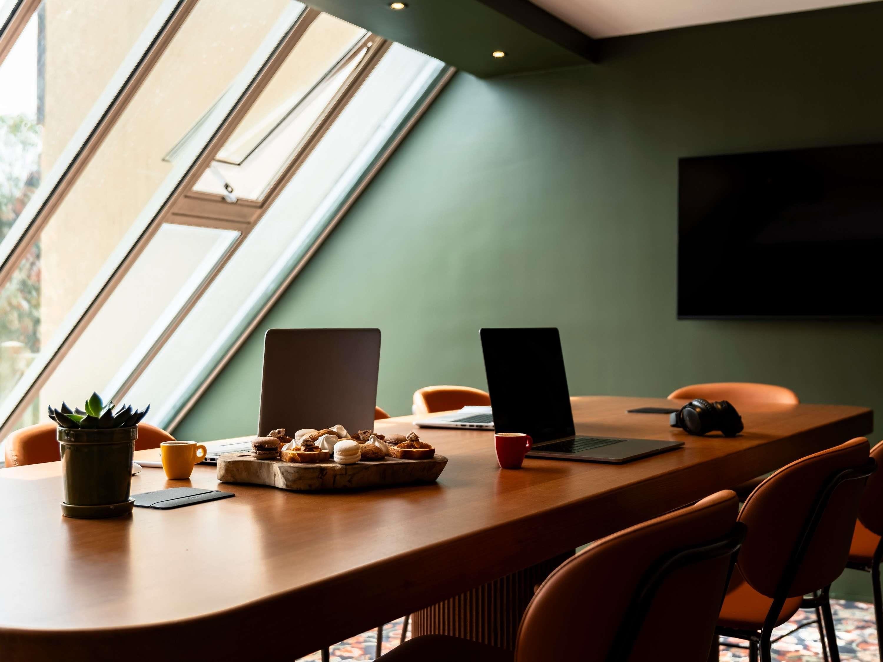A modern meeting room with a large table, two laptops, a plant, a tray of pastries, and a TV screen.