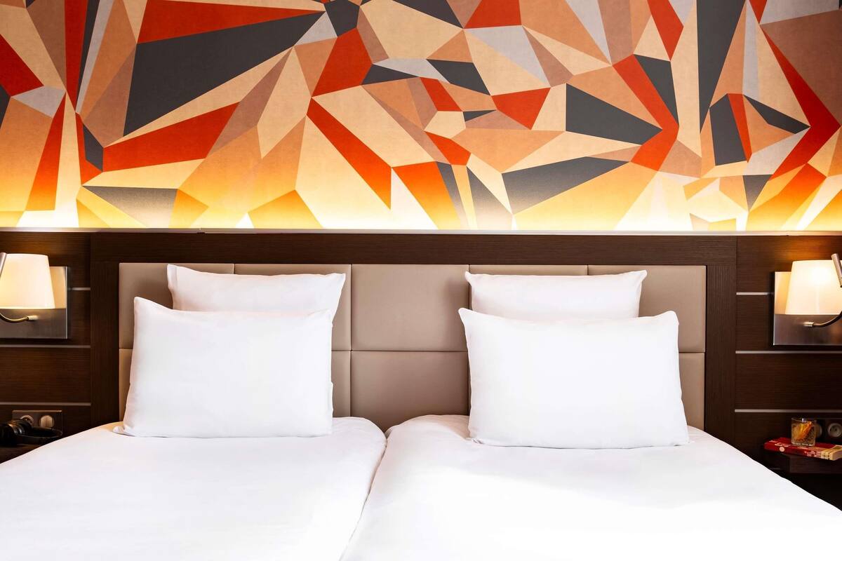 A hotel room with a modern geometric wall design, a tufted headboard, and two white beds with matching pillows.