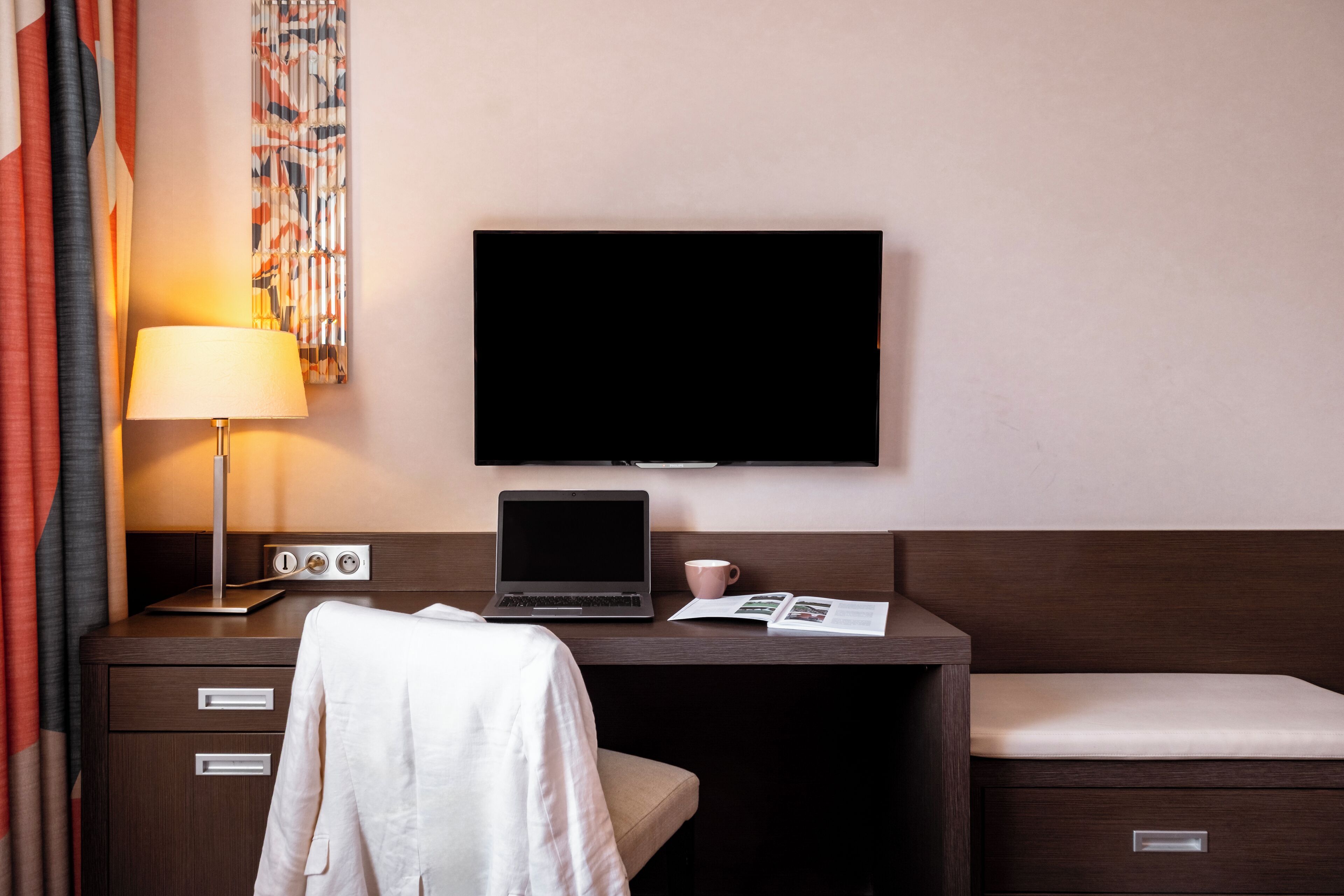 Ein Hotelzimmer mit einem Bett, einem Schreibtisch mit einem Laptop, einem Fernseher, einer Lampe und einem Fenster mit Vorhängen.