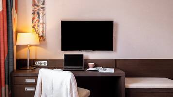 Ein Hotelzimmer mit einem Bett, einem Schreibtisch mit einem Laptop, einem Fernseher, einer Lampe und einem Fenster mit Vorhängen.