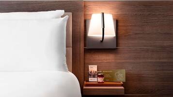 Habitación de hotel con una cama bien hecha, una lámpara de noche y una pequeña mesita de madera con un libro y una botella.