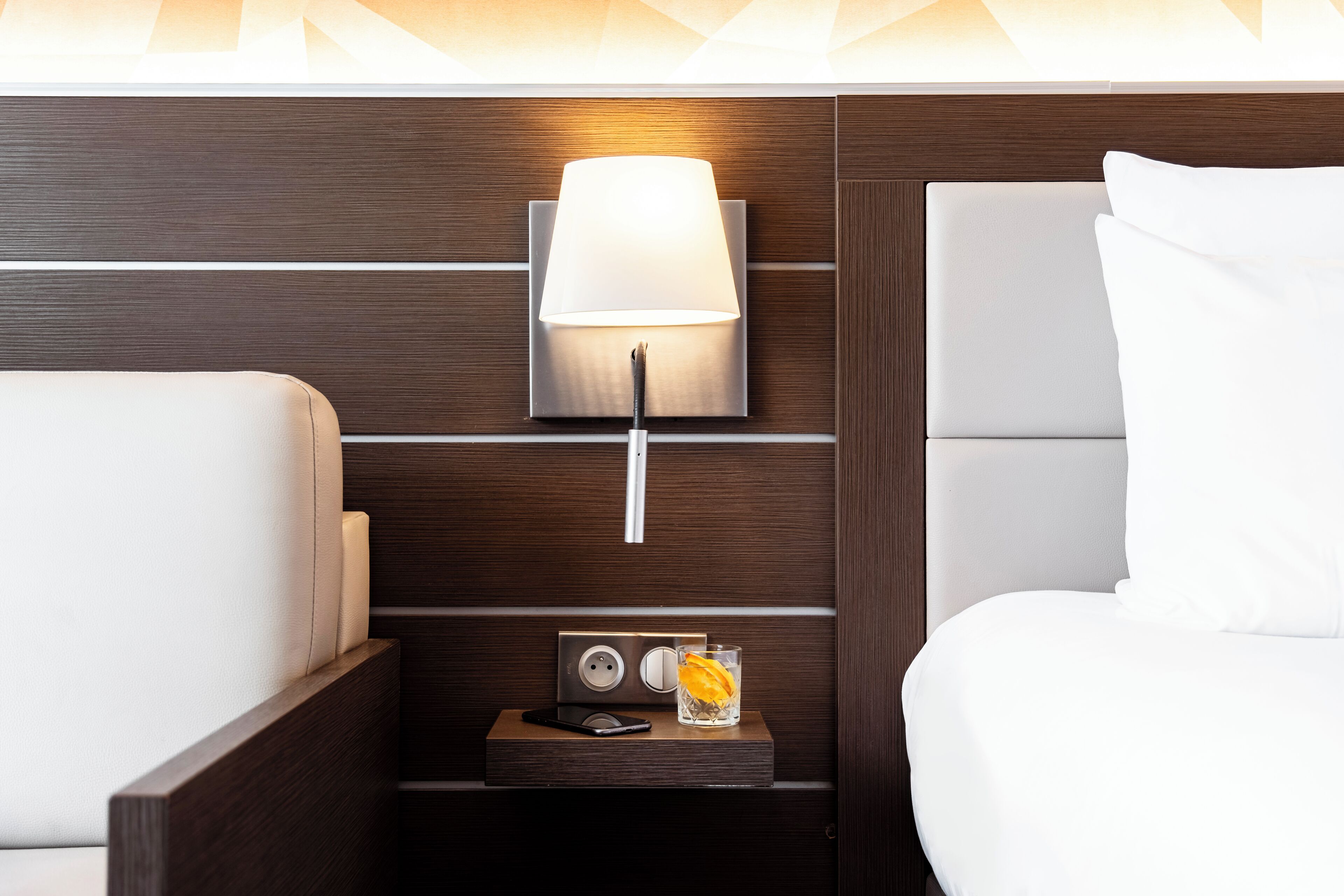 A neatly arranged hotel room with a modern design, featuring a bedside table with a lamp, a glass of water, and a small electronic device.