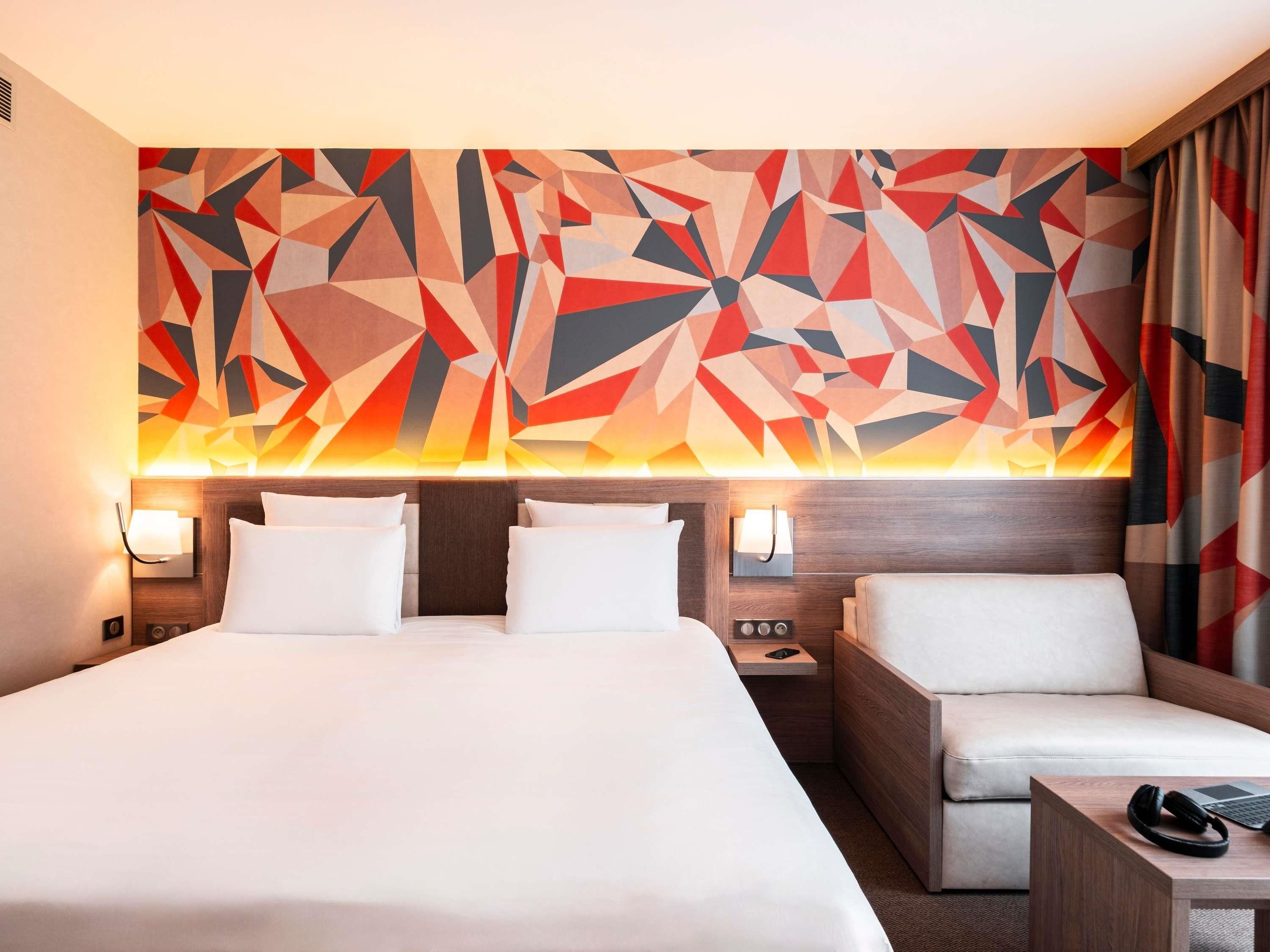 A hotel room with a large bed, two bedside tables, a sofa, and a geometric wall mural.