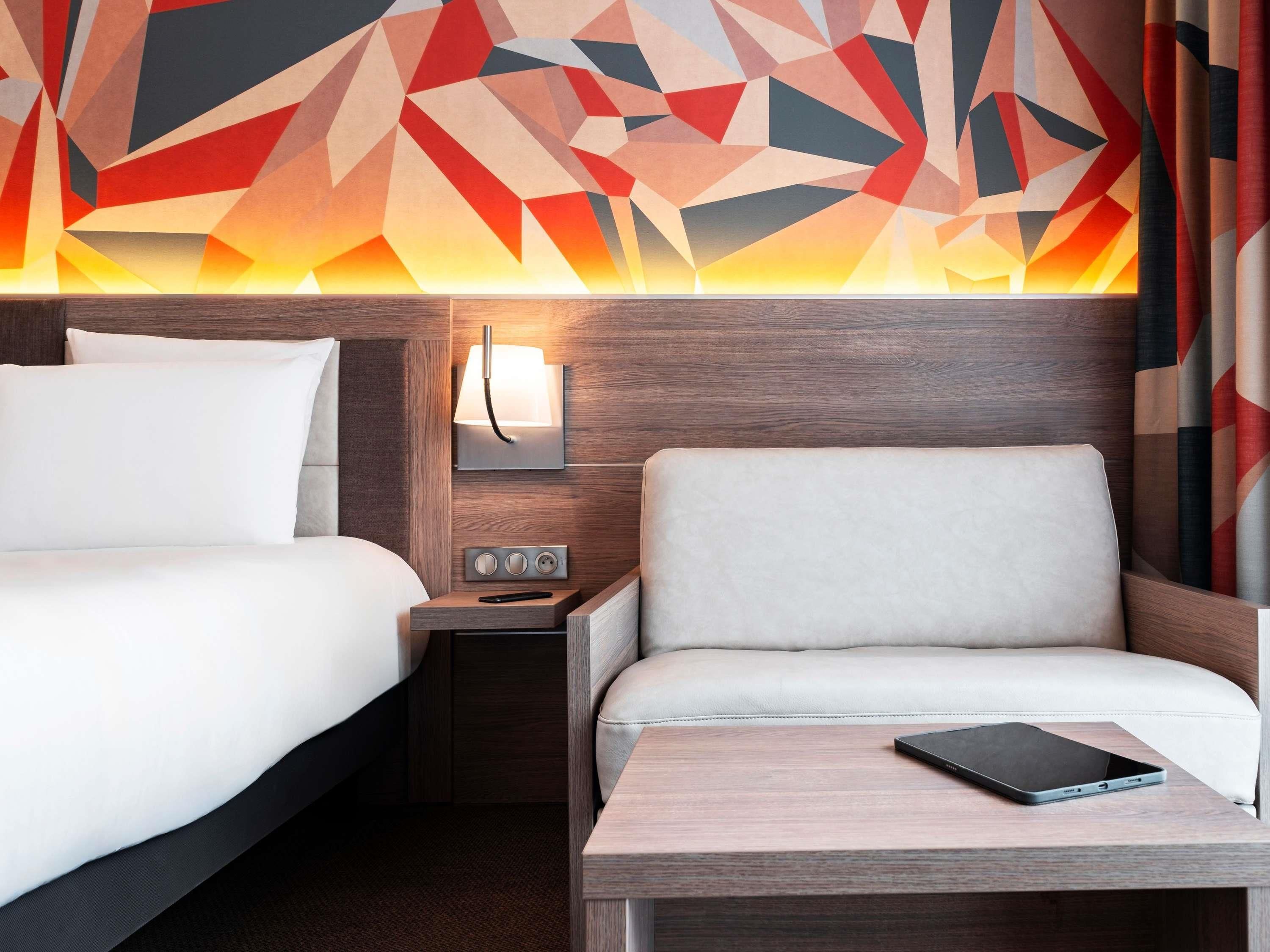 A hotel room with a bed, a bedside table with a lamp, a chair, and a wall with geometric patterns.