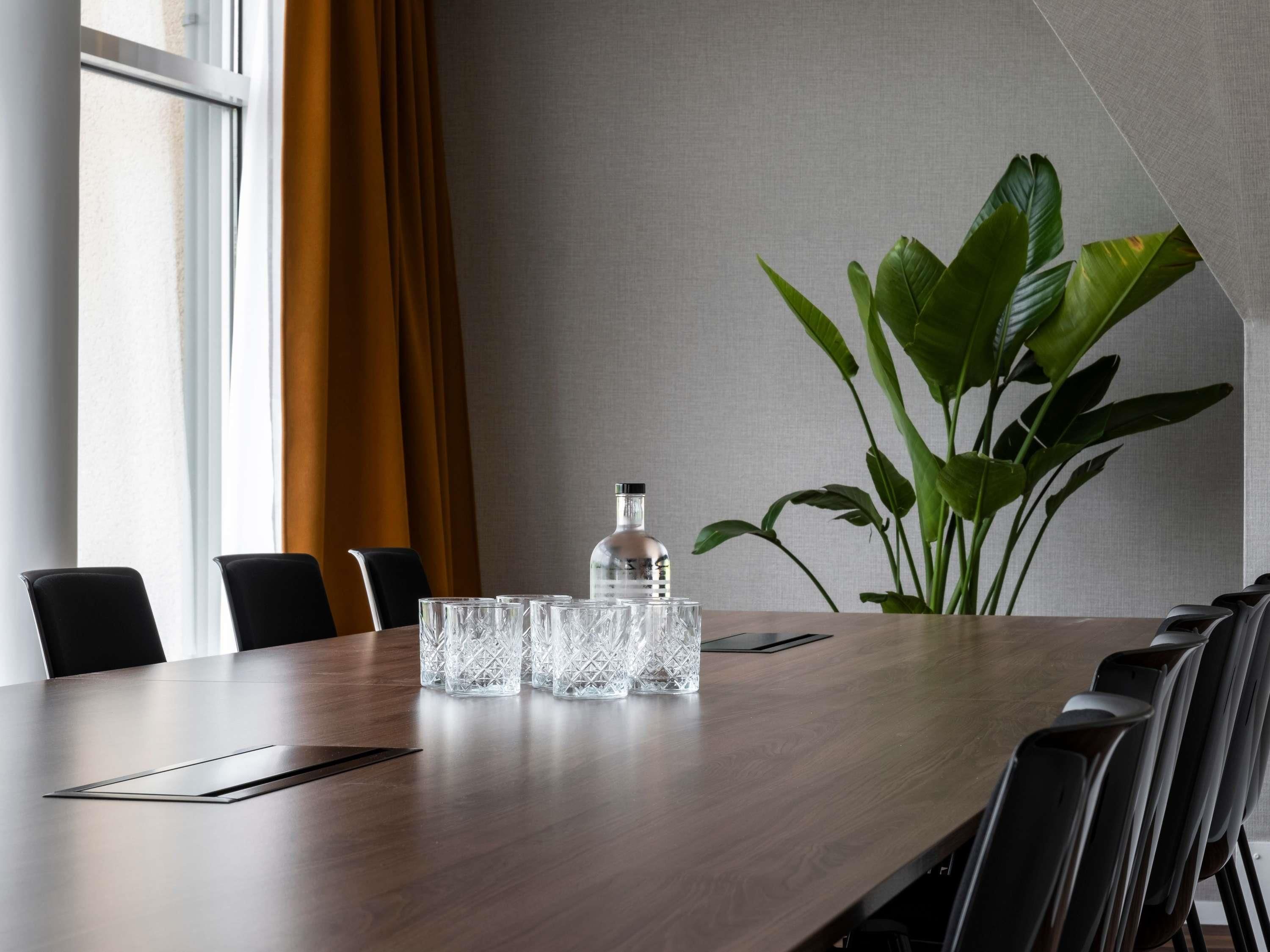 A conference table with chairs, a bottle, and glasses, with a plant and curtains in the background.