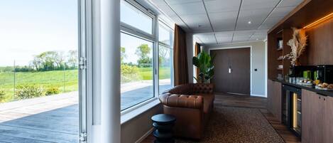 A modern interior with a large sliding glass door, a leather sofa, a kitchen area, and a view of a green landscape.