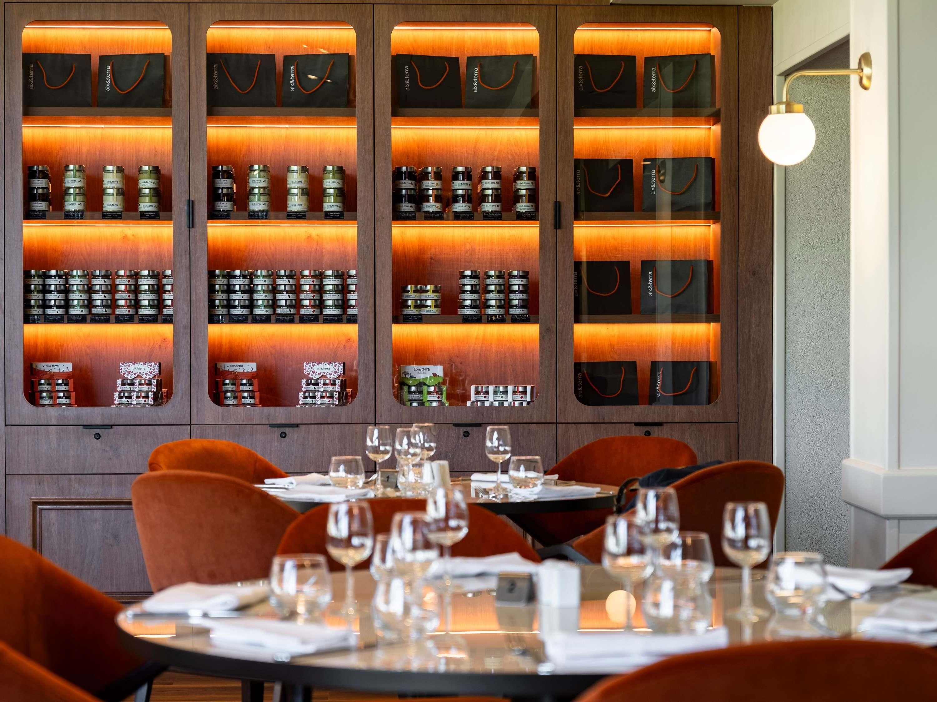 Un restaurante con un aparador de madera que exhibe diversas botellas, mesas dispuestas con copas de vino y sillas color naranja.