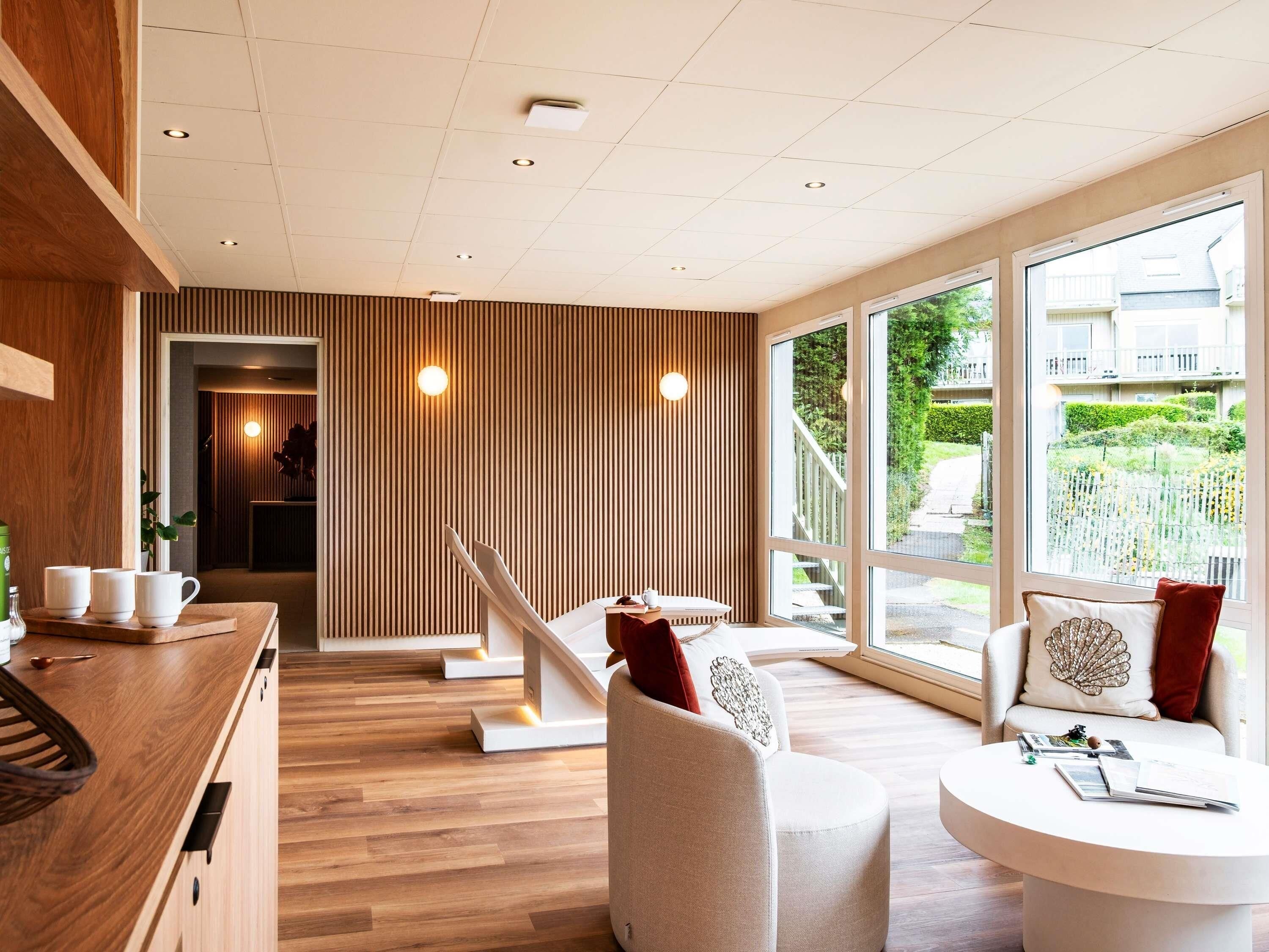 Una habitación de hotel moderna con suelo de madera, un sofá blanco, una mesa redonda y ventanas amplias con vistas al exterior.
